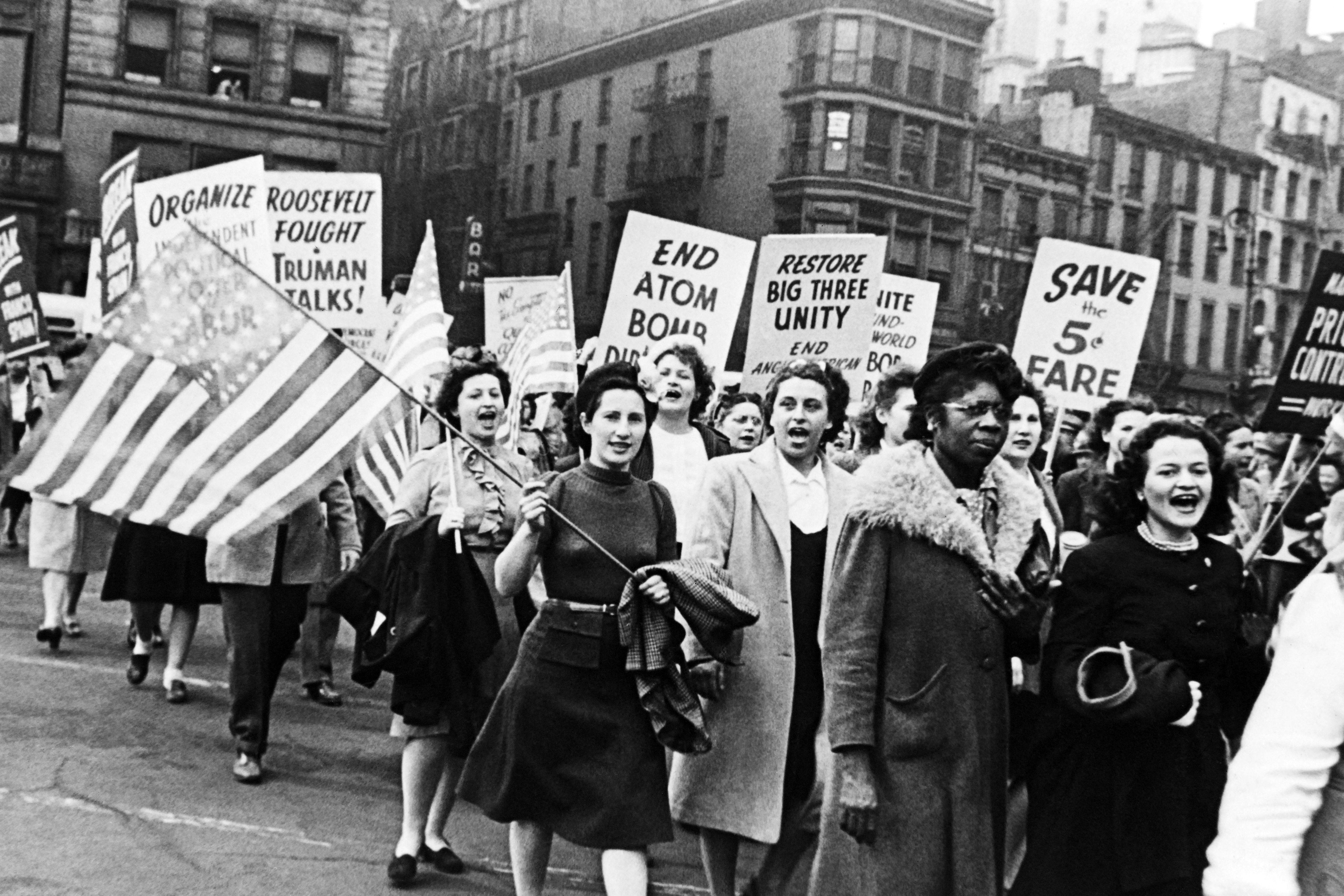 Women marching with protestation  signs advocating for atomic  disarmament and fare savings. They transportation  an American emblem  successful  a metropolis  setting