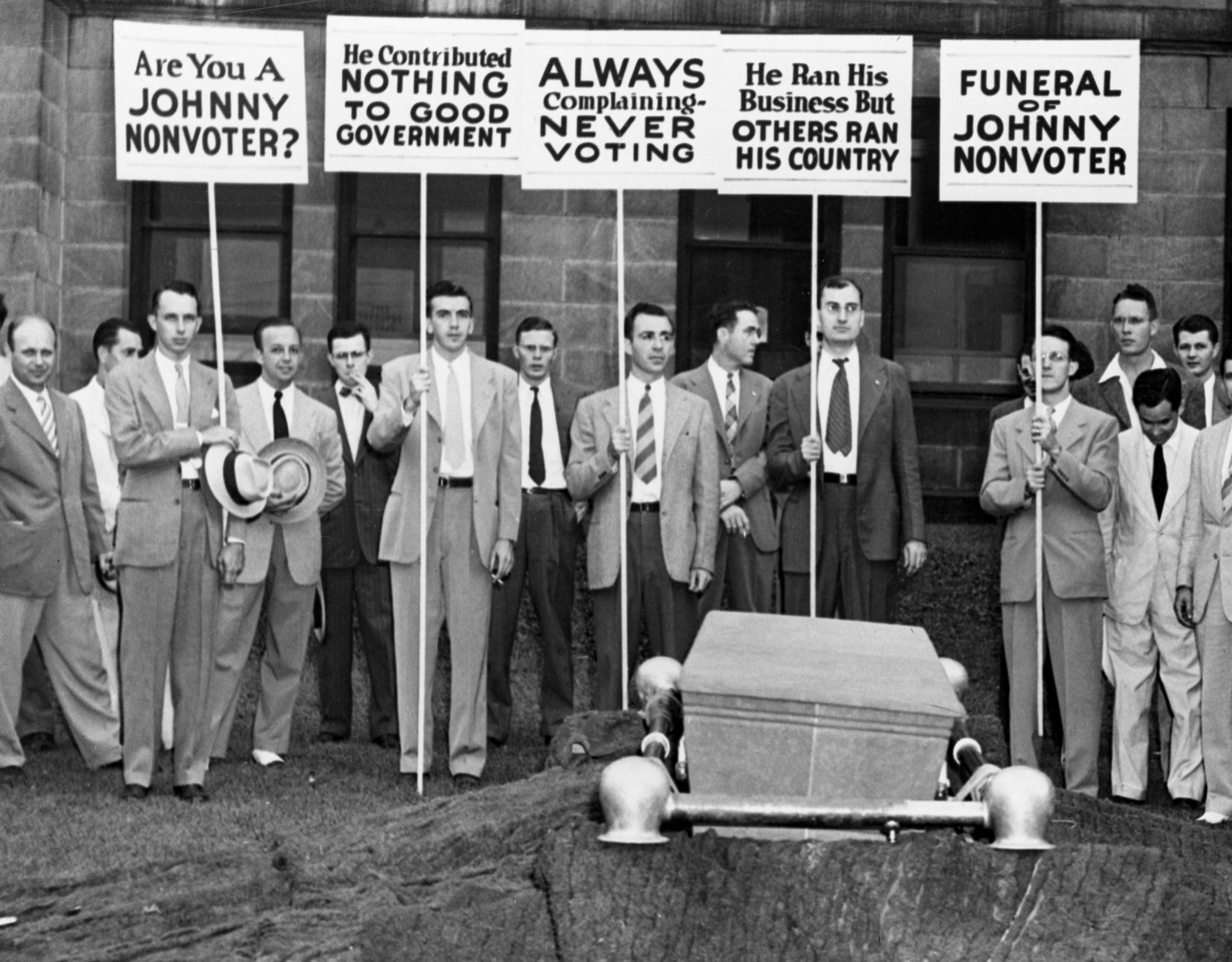 A radical  of men successful  suits holds protestation  signs astir  voting, surrounding a coffin labeled "Funeral of Johnny Nonvoter."