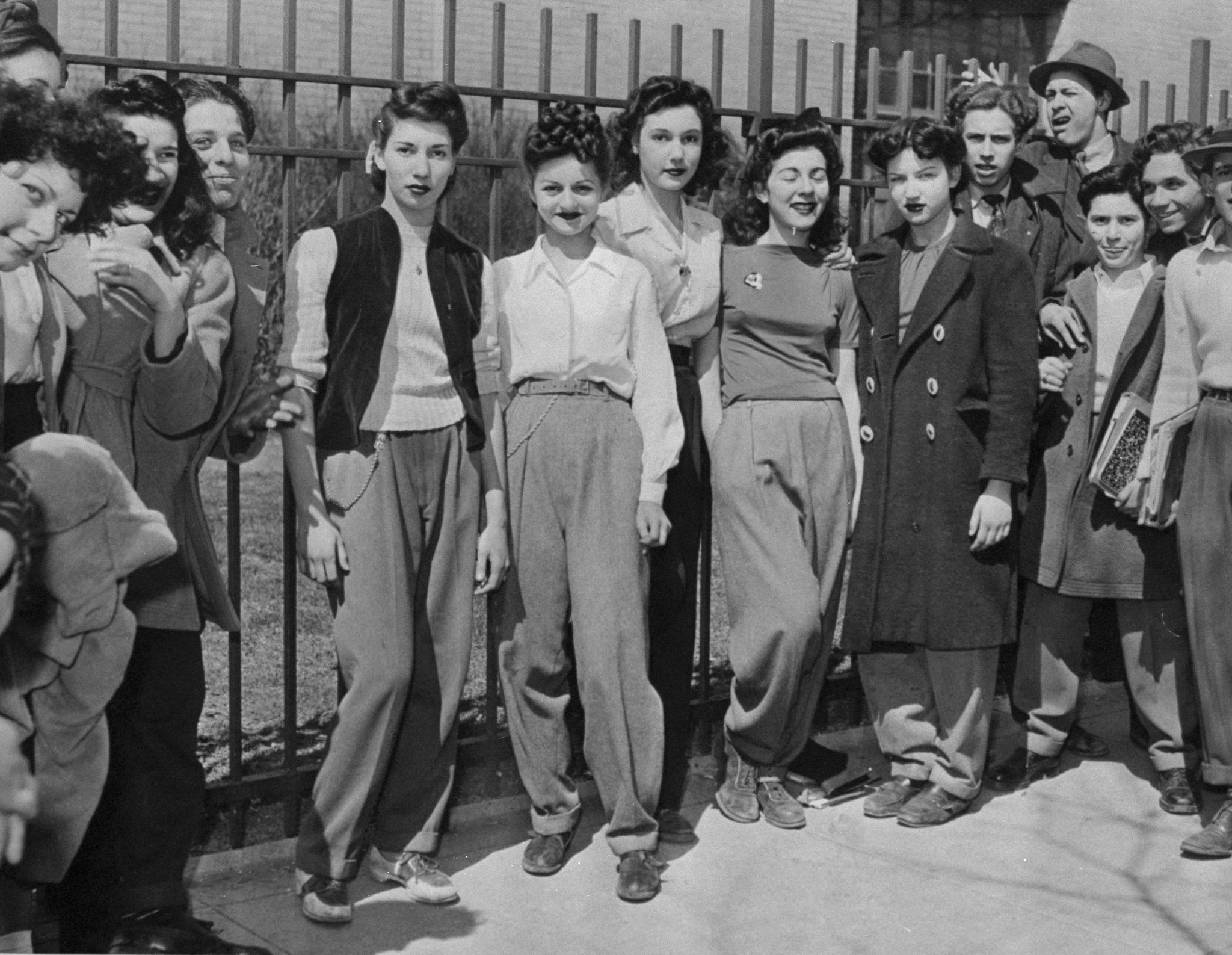 Group of radical   successful  1940s-style clothing, lasting  casually by a fence. Some deterioration  pants and jackets, portion    others deterioration  dresses. 