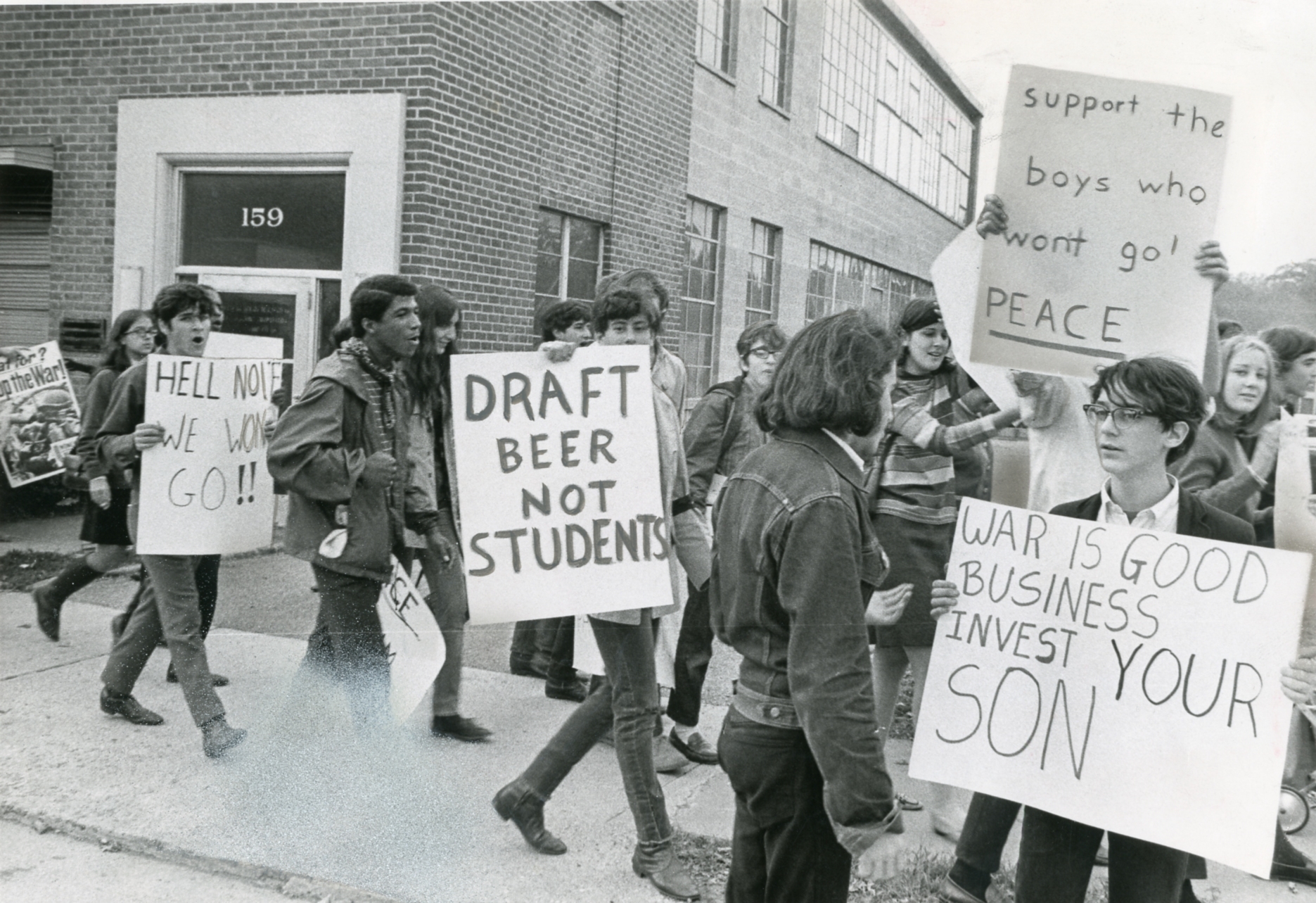 Protesters clasp  signs with anti-war and anti-draft messages extracurricular  a building