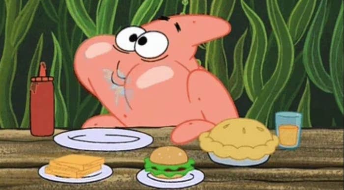 Animated character Patrick Star eagerly looks at a meal with sandwiches, a burger, pie, and a drink on a wooden table
