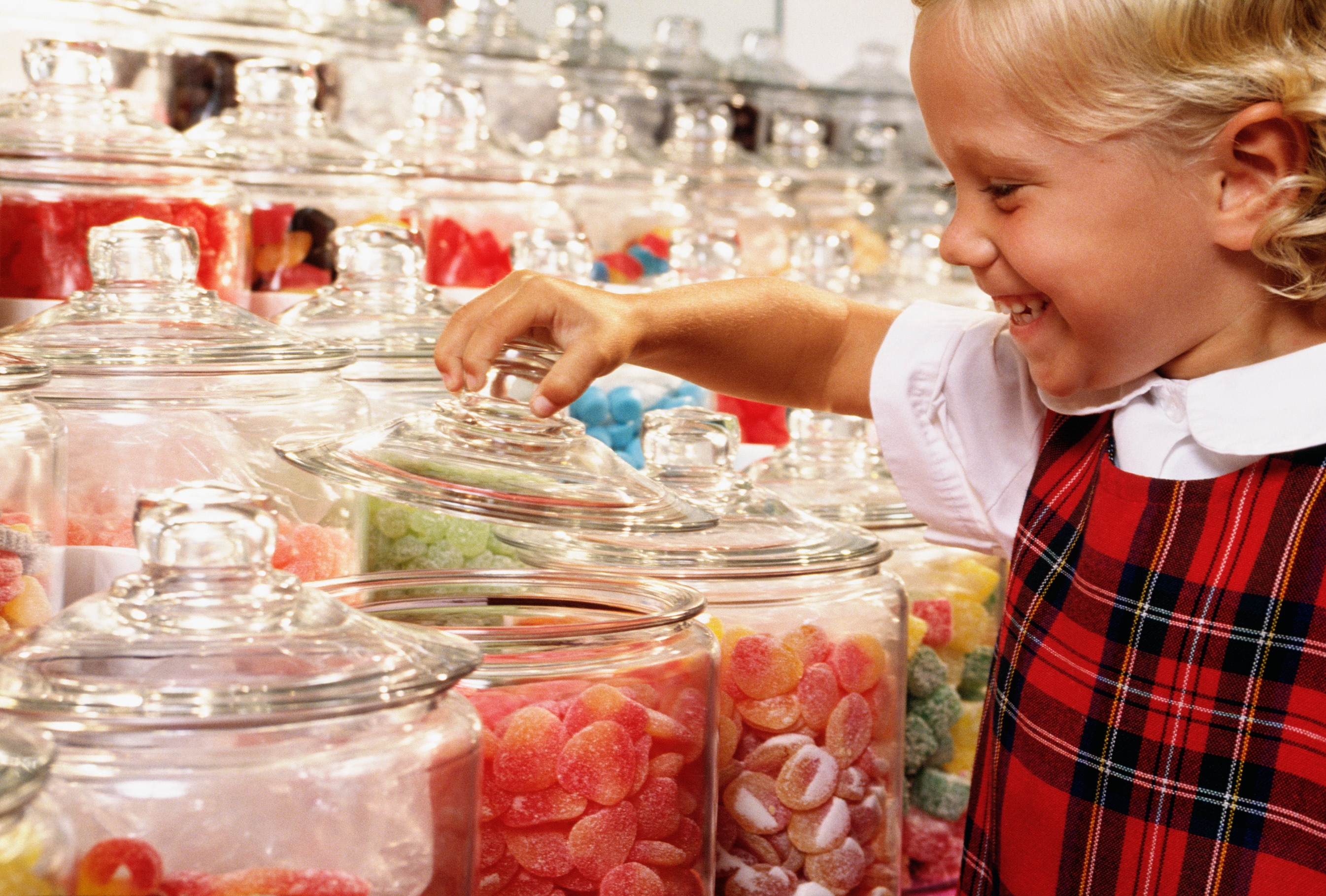 Child smiling while selecting candy from large glass jars in a store. The child is wearing a plaid dress