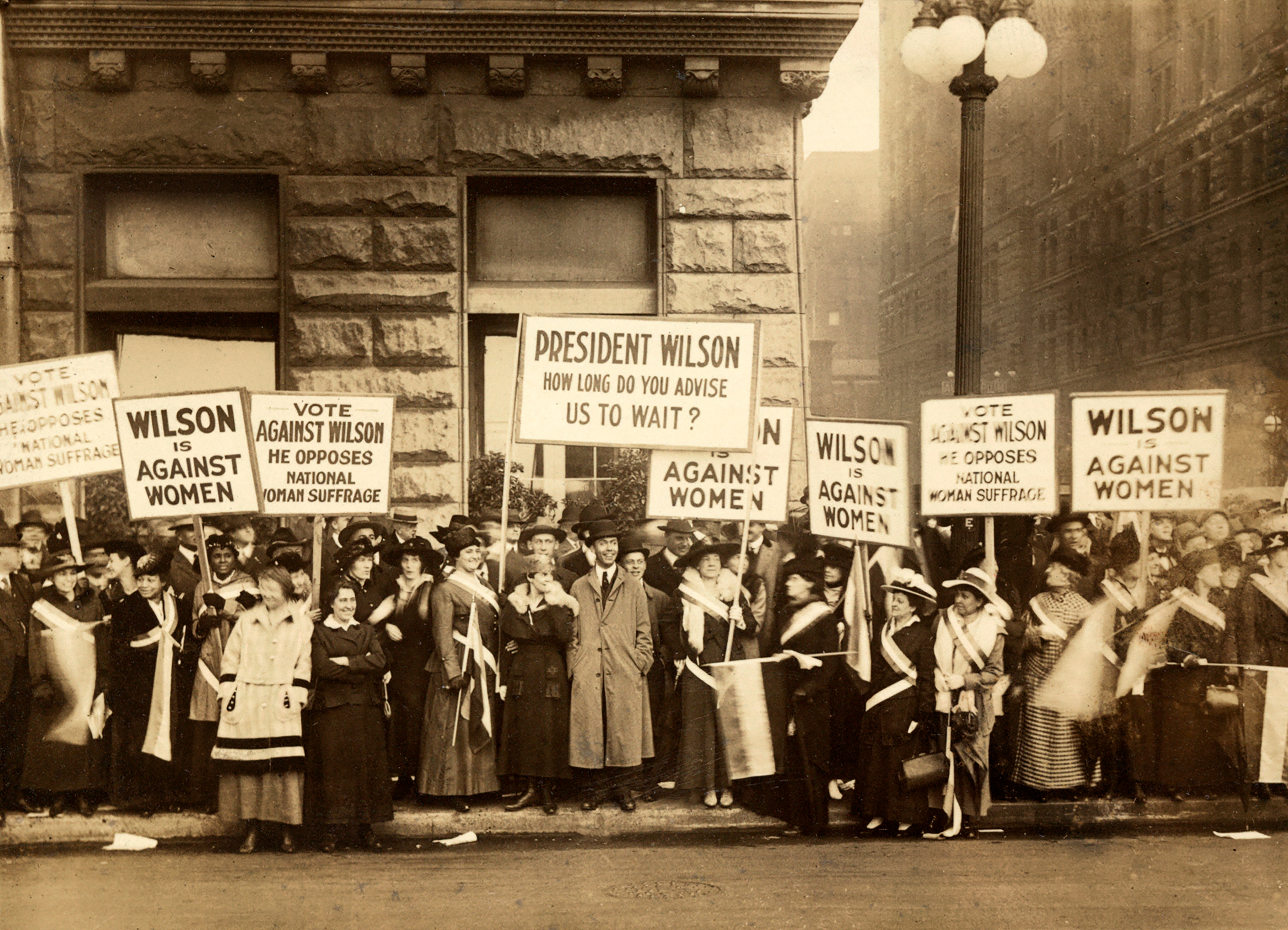Historical protestation  with women holding signs demanding voting rights and criticizing President Wilson's stance against women's suffrage