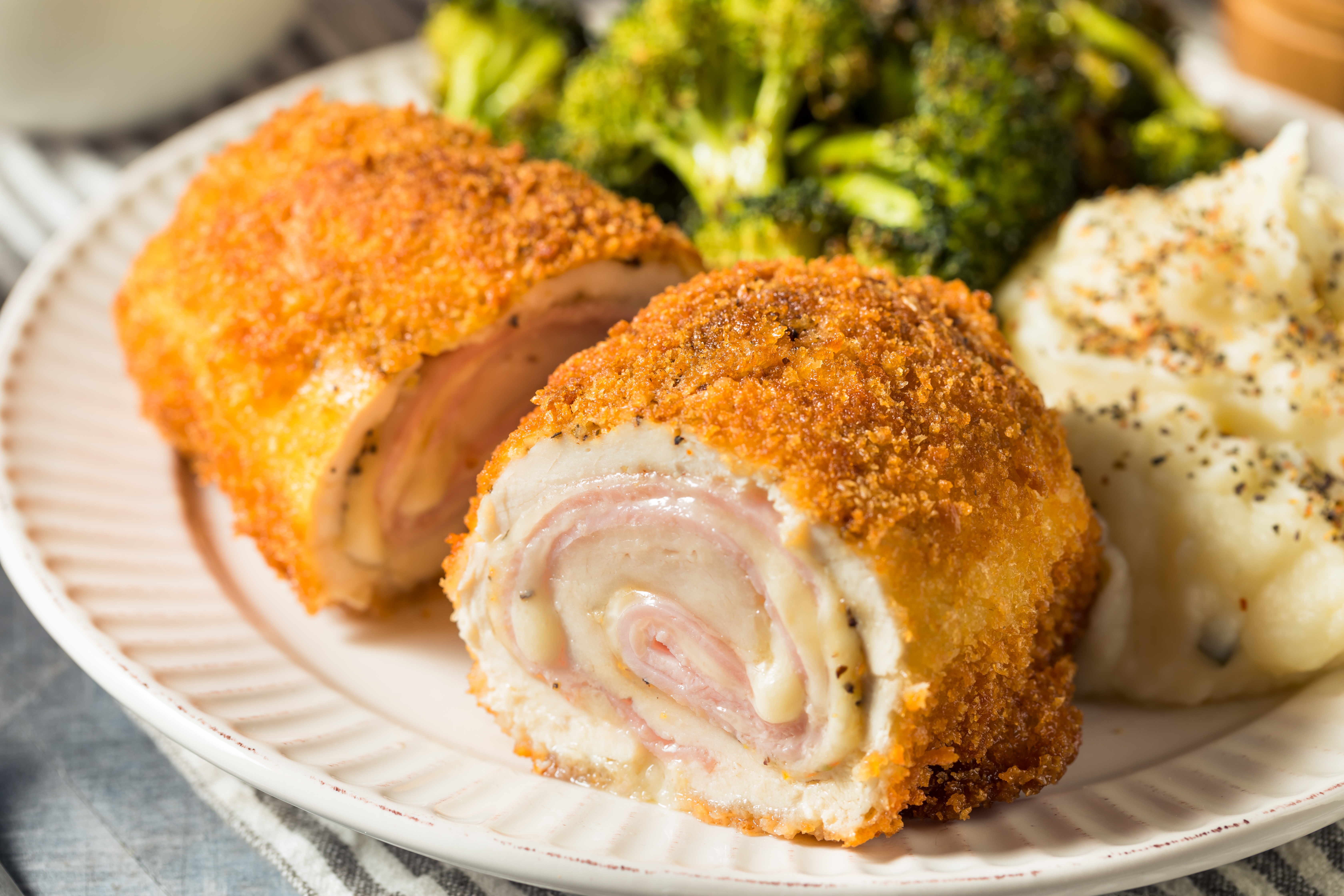 Close-up of chicken cordon bleu on a plate with mashed potatoes and broccoli, showcasing its crispy breading and layered ham and cheese inside