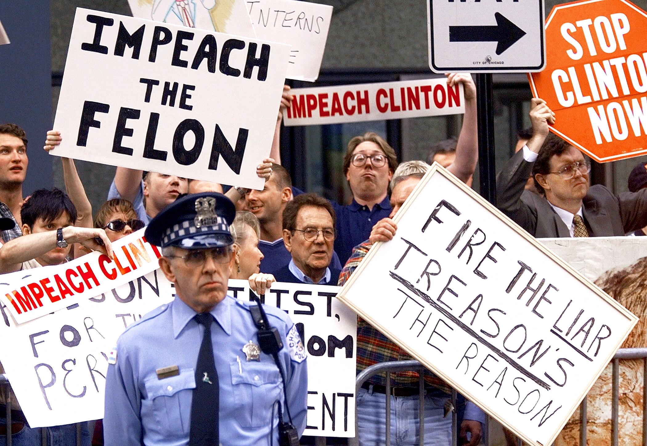 Protesters holding signs against Clinton, including "Impeach the Felon" and "Fire the Liar." A constabulary  serviceman  stands successful  front