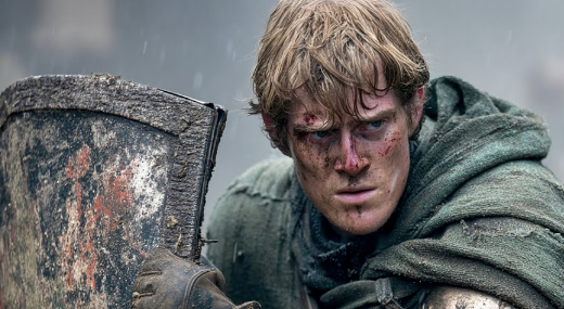 Actor in a rugged medieval costume, holding a shield, with a determined expression and battle-worn face, suggesting a scene from a period drama