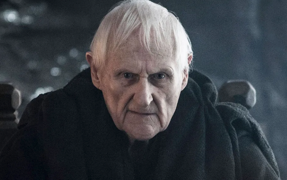 Elderly person with white hair in dark robe, sitting solemnly, exudes a serious expression in a dramatic, shadowed setting