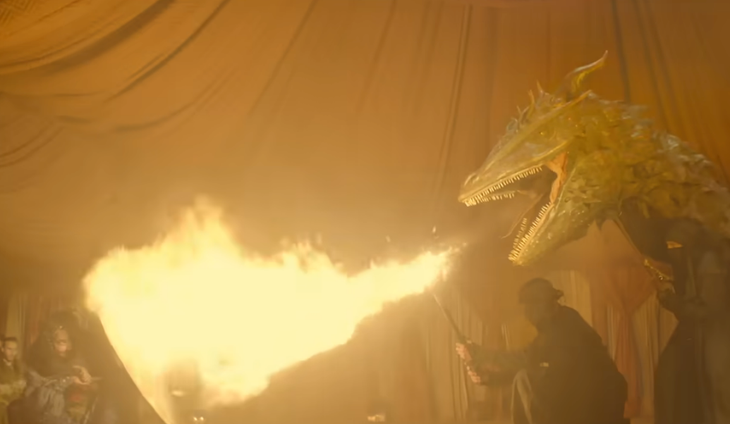 A dragon breathes fire inside a large tent as a person holds it on a leash, evoking a fantasy scene
