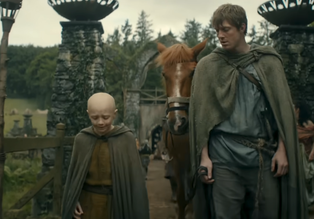 Two characters in medieval cloaks walk with a horse through a stone gate in a rustic, historical setting, suggesting a fantasy storyline