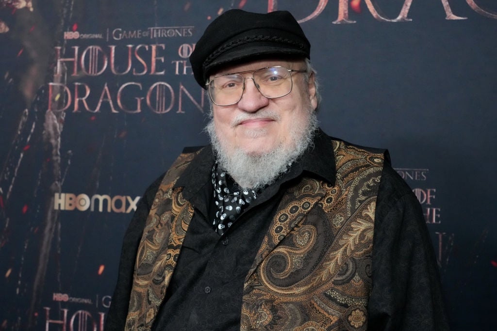 A person wearing a patterned vest, black shirt, and cap stands in front of a "House of the Dragon" backdrop at a TV event