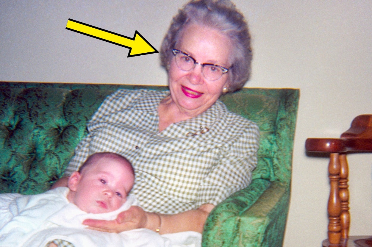 Elderly woman with glasses, seated on a green couch, holding a baby in her lap. Yellow arrow pointing at her