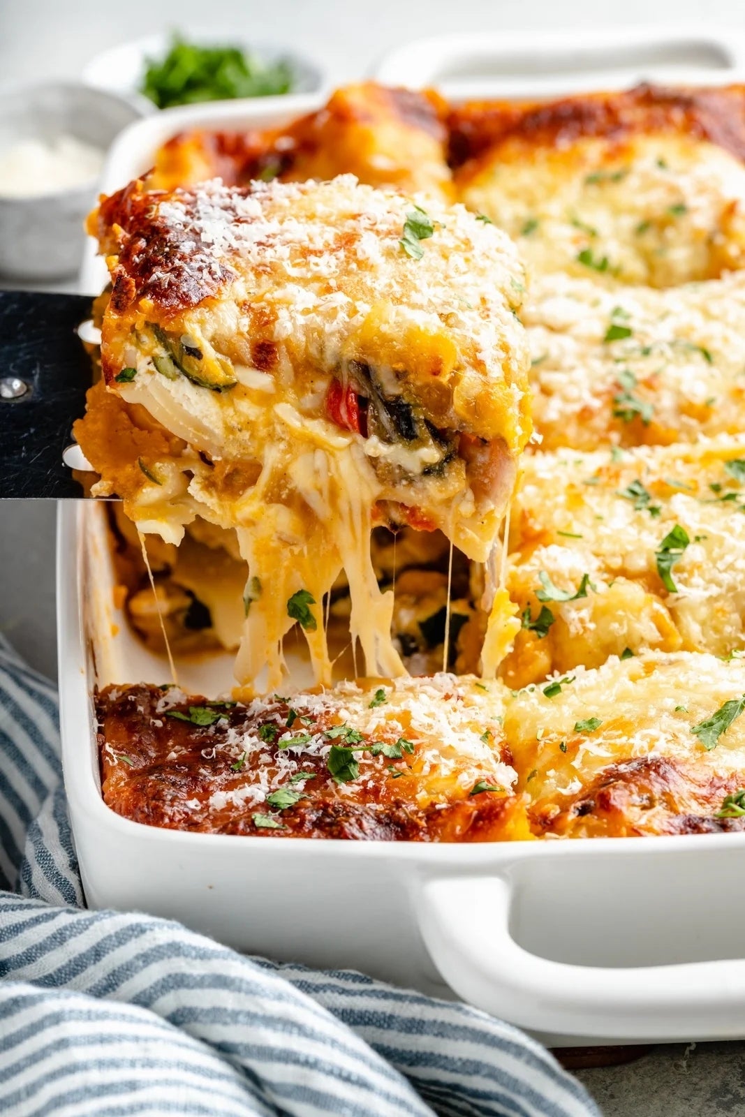 A cheesy, baked lasagna being served from a dish, with layers of pasta, vegetables, and cheese, garnished with herbs