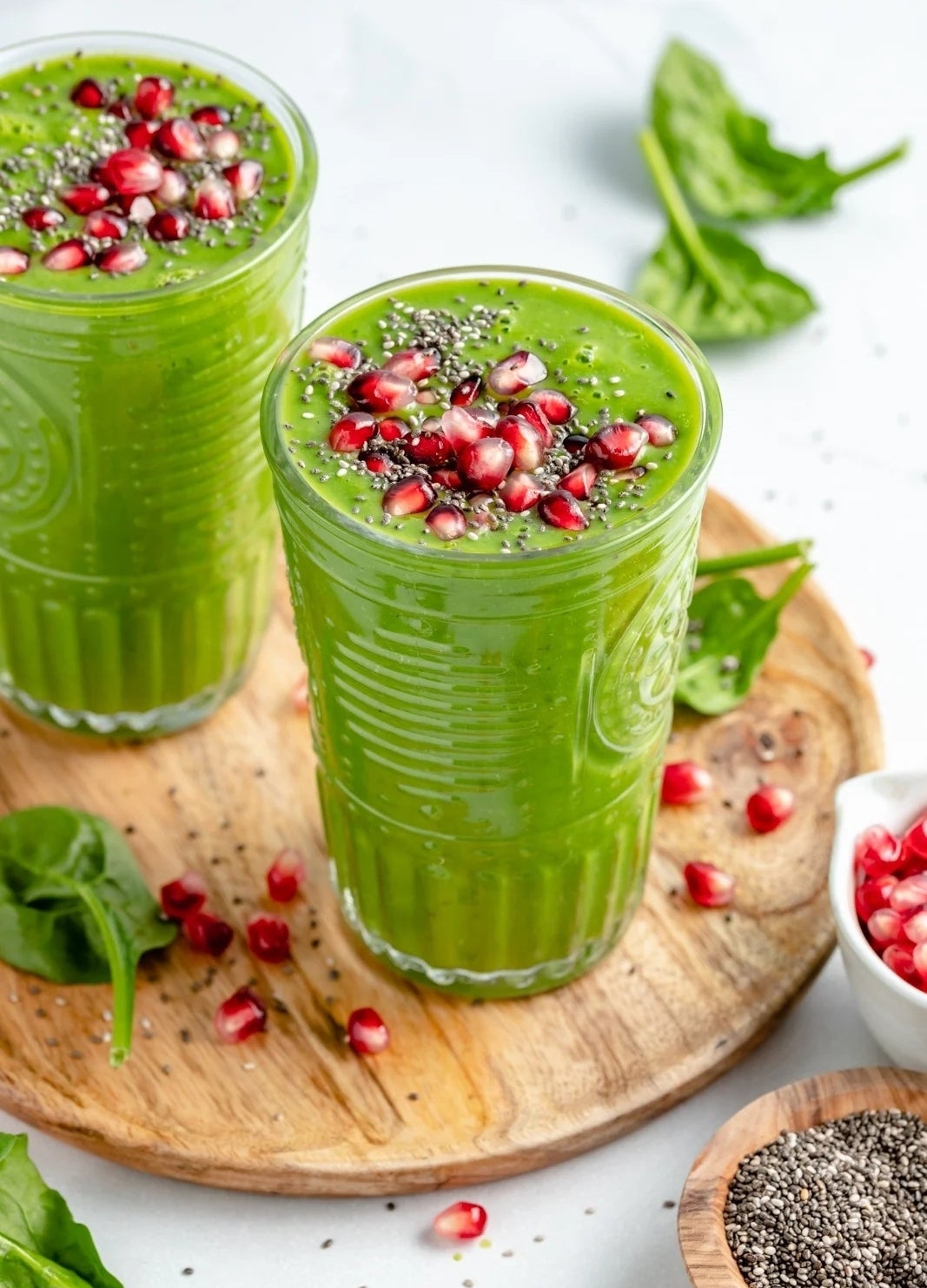 Two glasses of green smoothie topped with chia seeds and pomegranate arils, surrounded by spinach leaves and more pomegranate on a wooden board