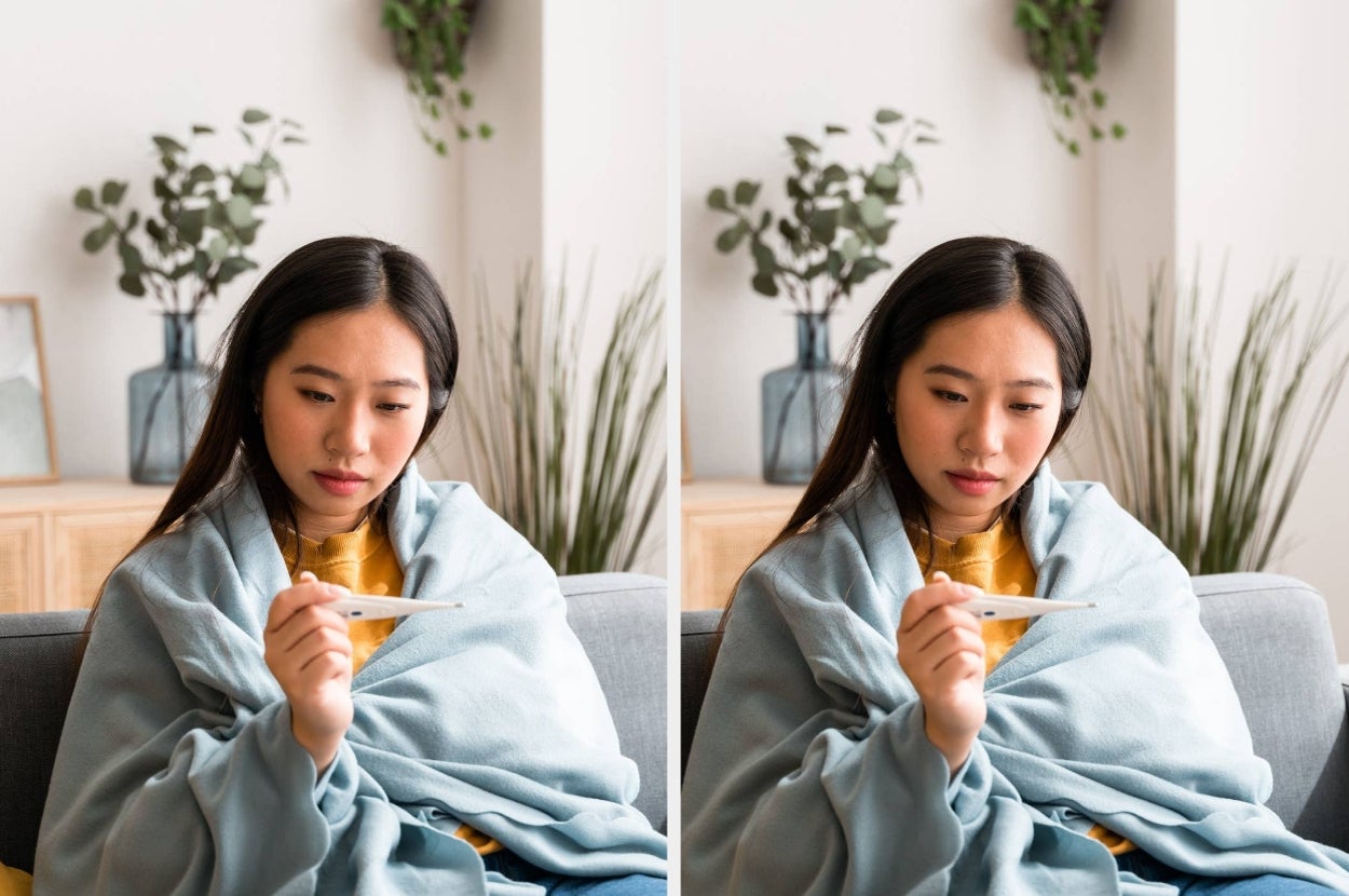 Woman on a couch under a blanket, looking at a thermometer with a thoughtful expression, suggesting she's checking her temperature