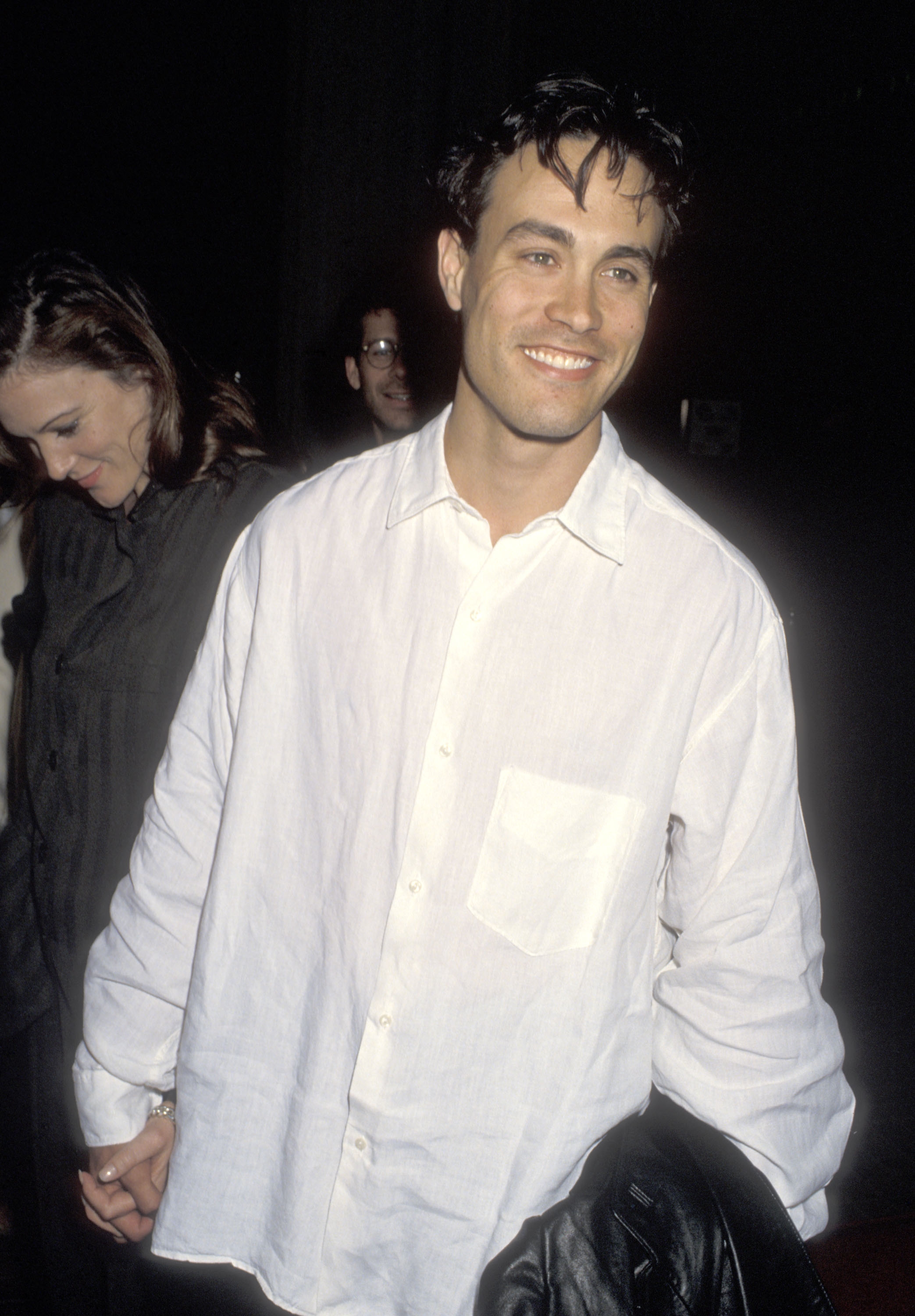 Person in a casual white shirt smiling, holding a jacket. A woman is partially visible beside them