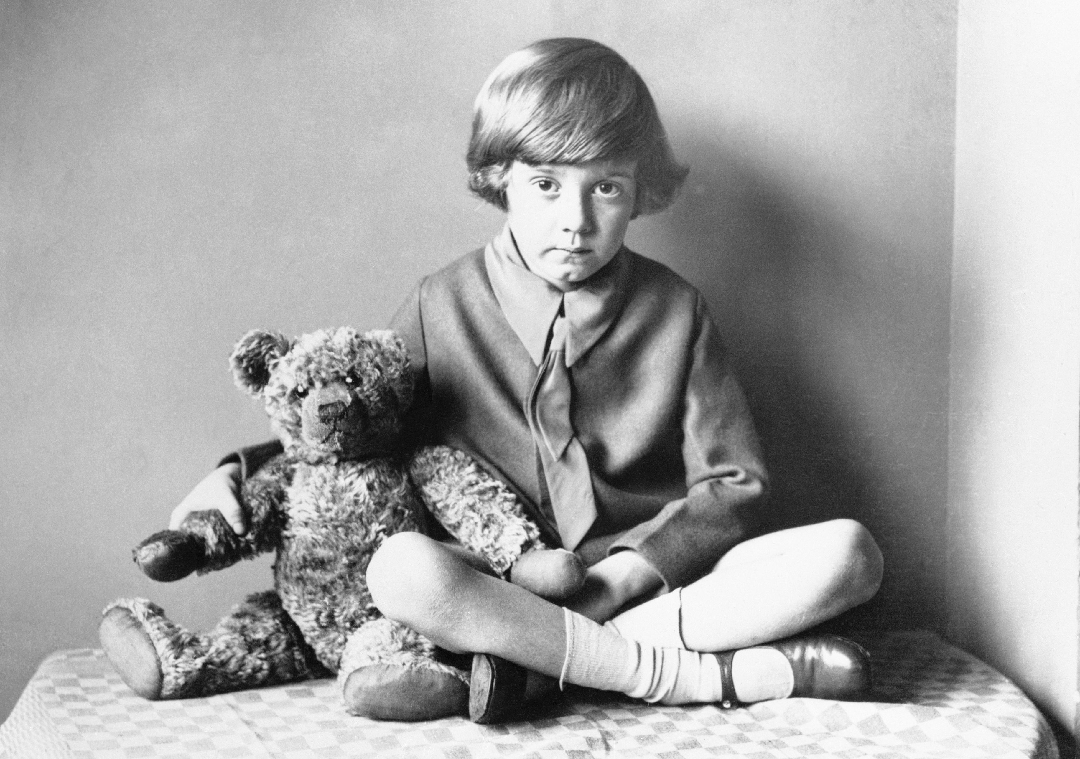 Child with 1920s-style haircut sits cross-legged beside a teddy bear, wearing a tie and sweater
