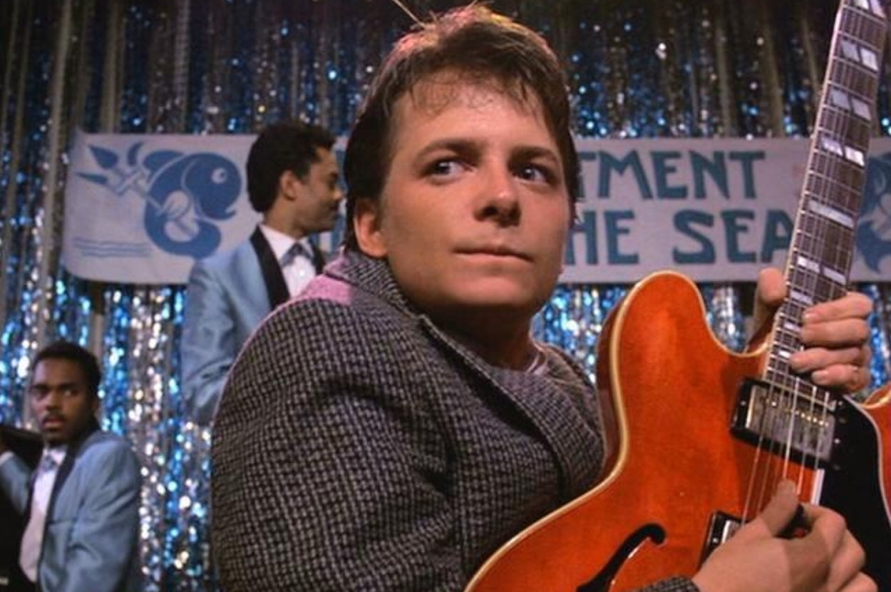 Person in a suit jacket holds a guitar on stage at an event with a "Sea" banner, while others are in the background