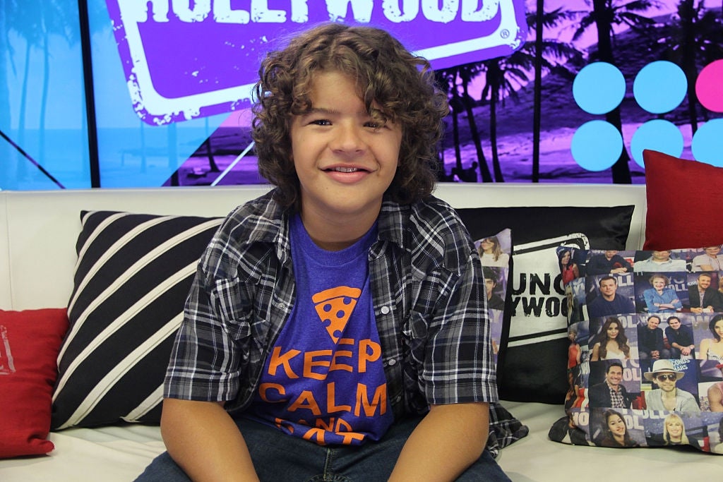 Seseorang dengan rambut keriting duduk di sofa mengenakan kemeja kotak-kotak di atas kaos bergambar. Latar belakangnya memiliki dekorasi bertema Hollywood