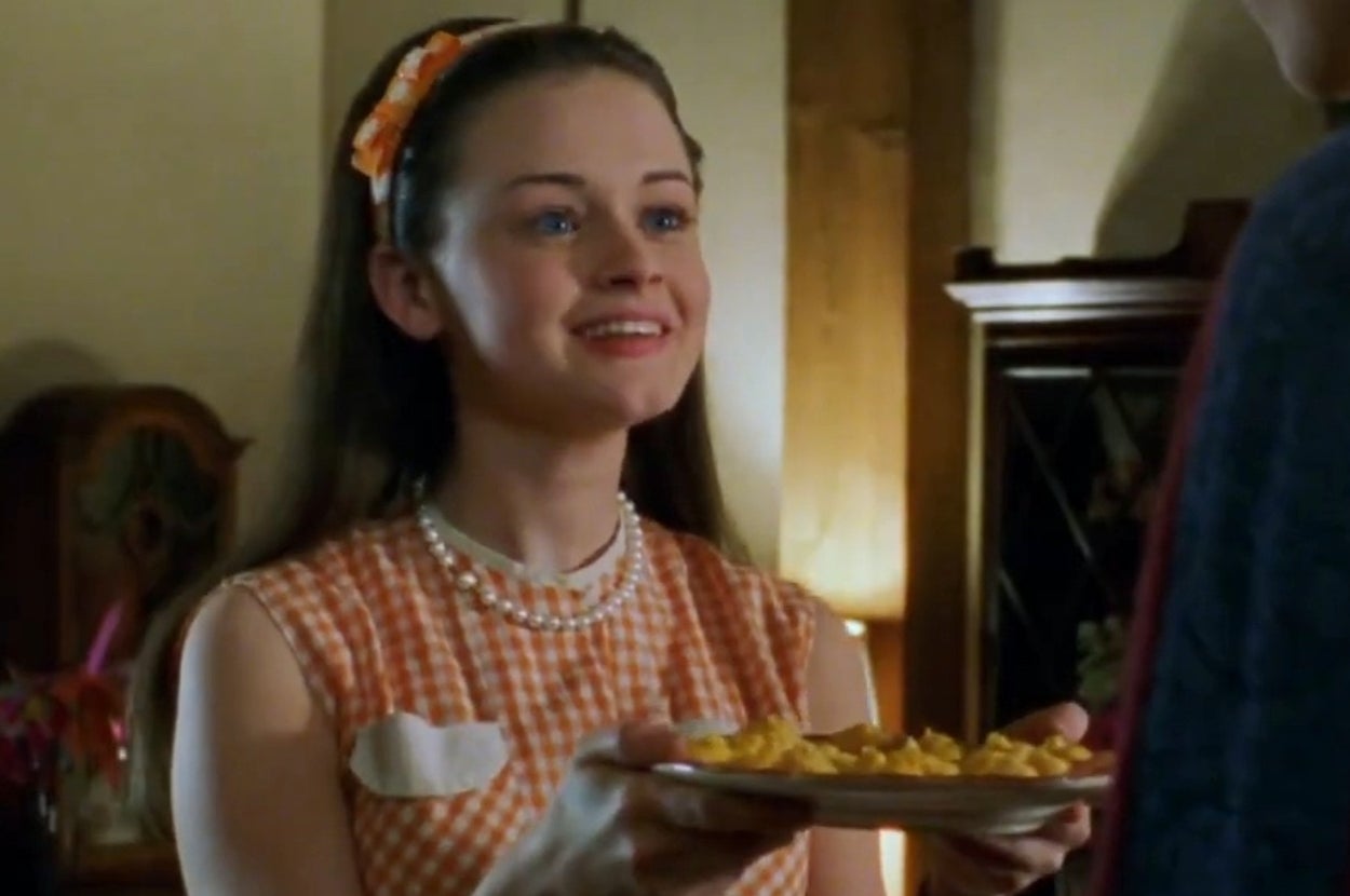 Person smiling and holding a plate of scrambled eggs, wearing a gingham dress with a headband, pearl necklace, and earrings, indoors