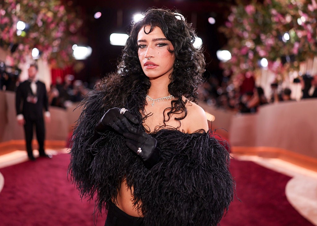 Seseorang dengan pakaian berbulu hitam yang elegan di karpet merah, berpose percaya diri dengan tampilan glamor, dikelilingi oleh fotografer dan dekorasi bunga