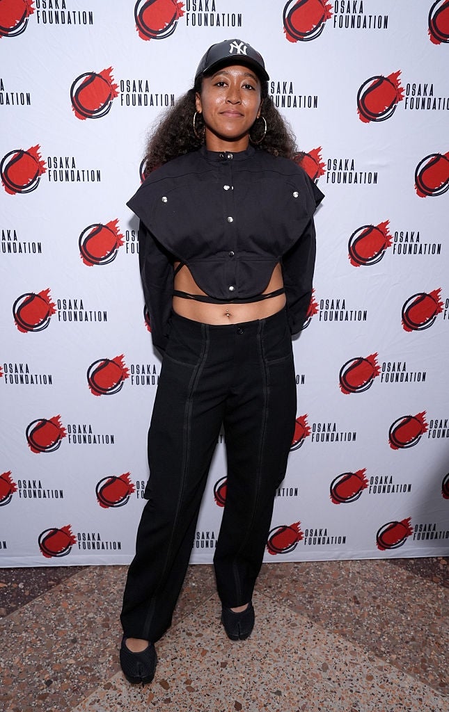 Seseorang dengan crop top dan celana hitam, mengenakan topi baseball, berpose di karpet merah di depan latar belakang berlogo Osaka Foundation