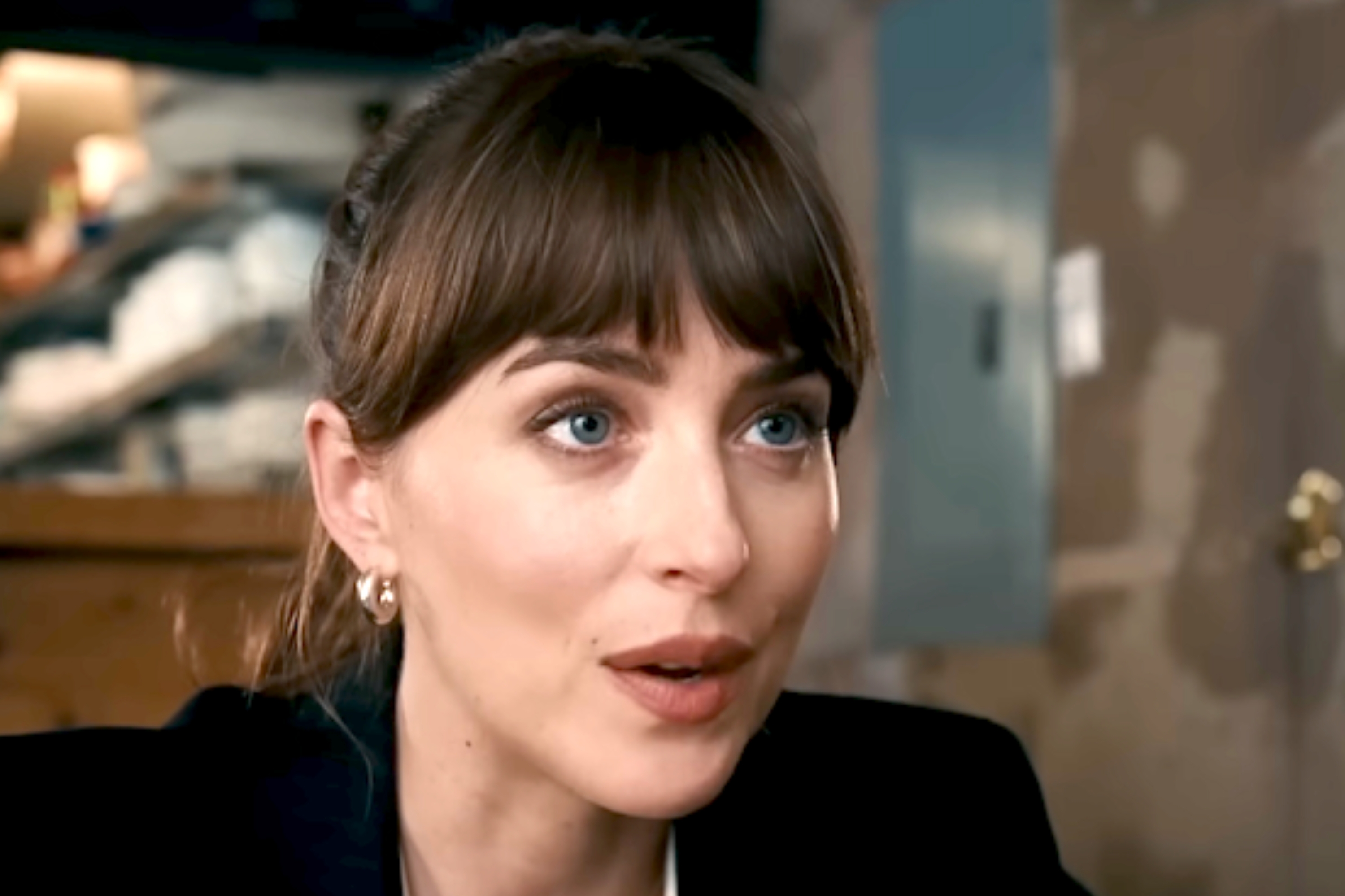 Person in a blazer sitting indoors, speaking during an interview or conversation. Background shows a rustic décor with shelves and a round mirror