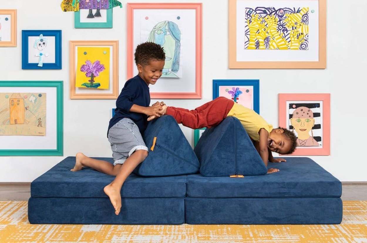 Two kids play on modular foam furniture blocks in a room with colorful framed children's artwork on the walls