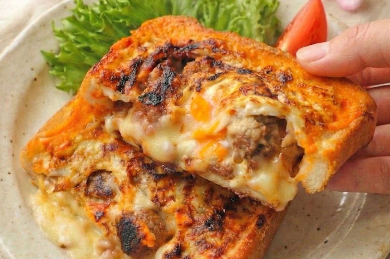 チーズとミートがたっぷりのトーストが皿にのり、サラダとトマトの付け合わせ。手がトーストを持ち上げている。