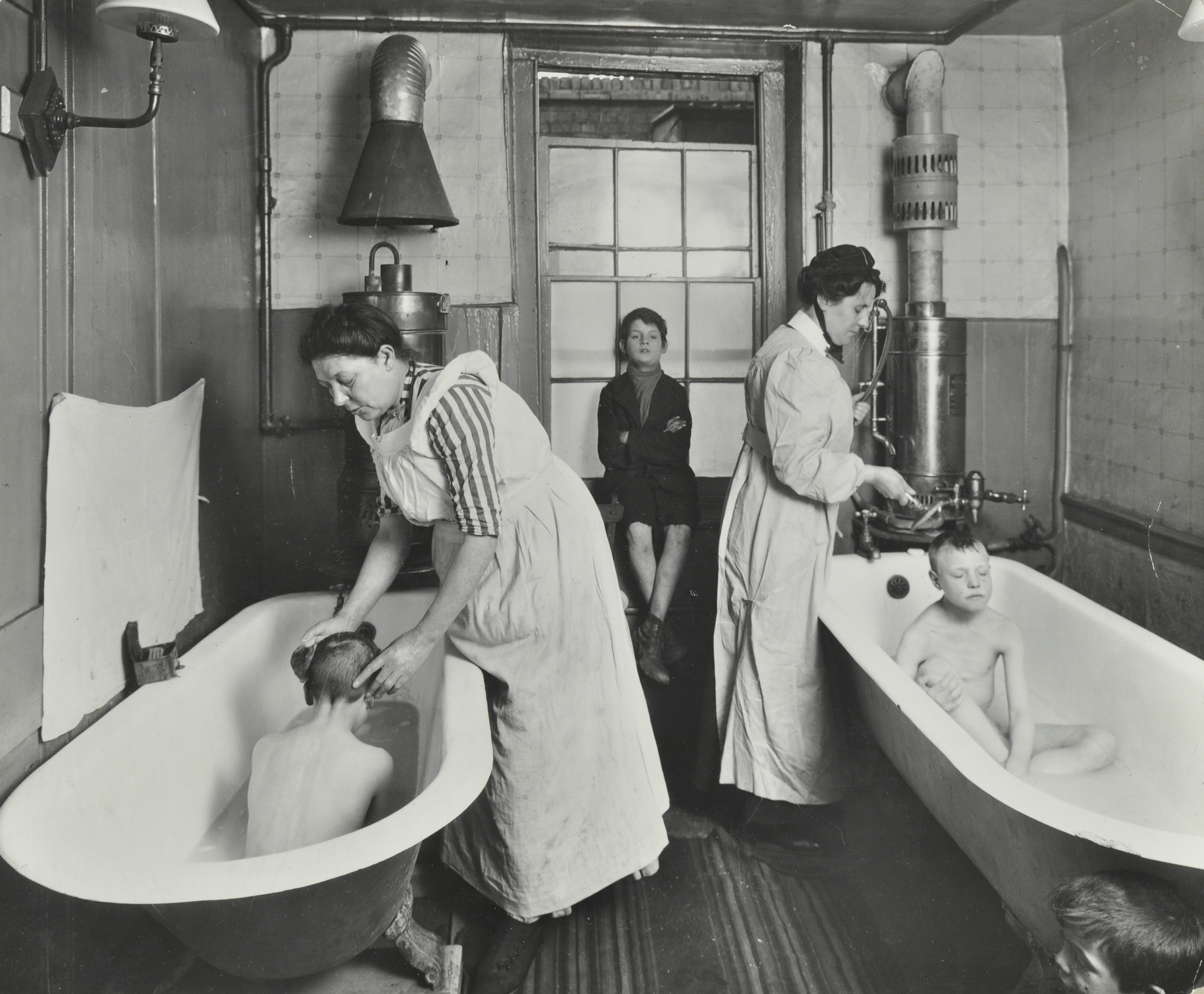 Two children are being bathed in separate bathtubs by women in uniforms, while another child sits in the background