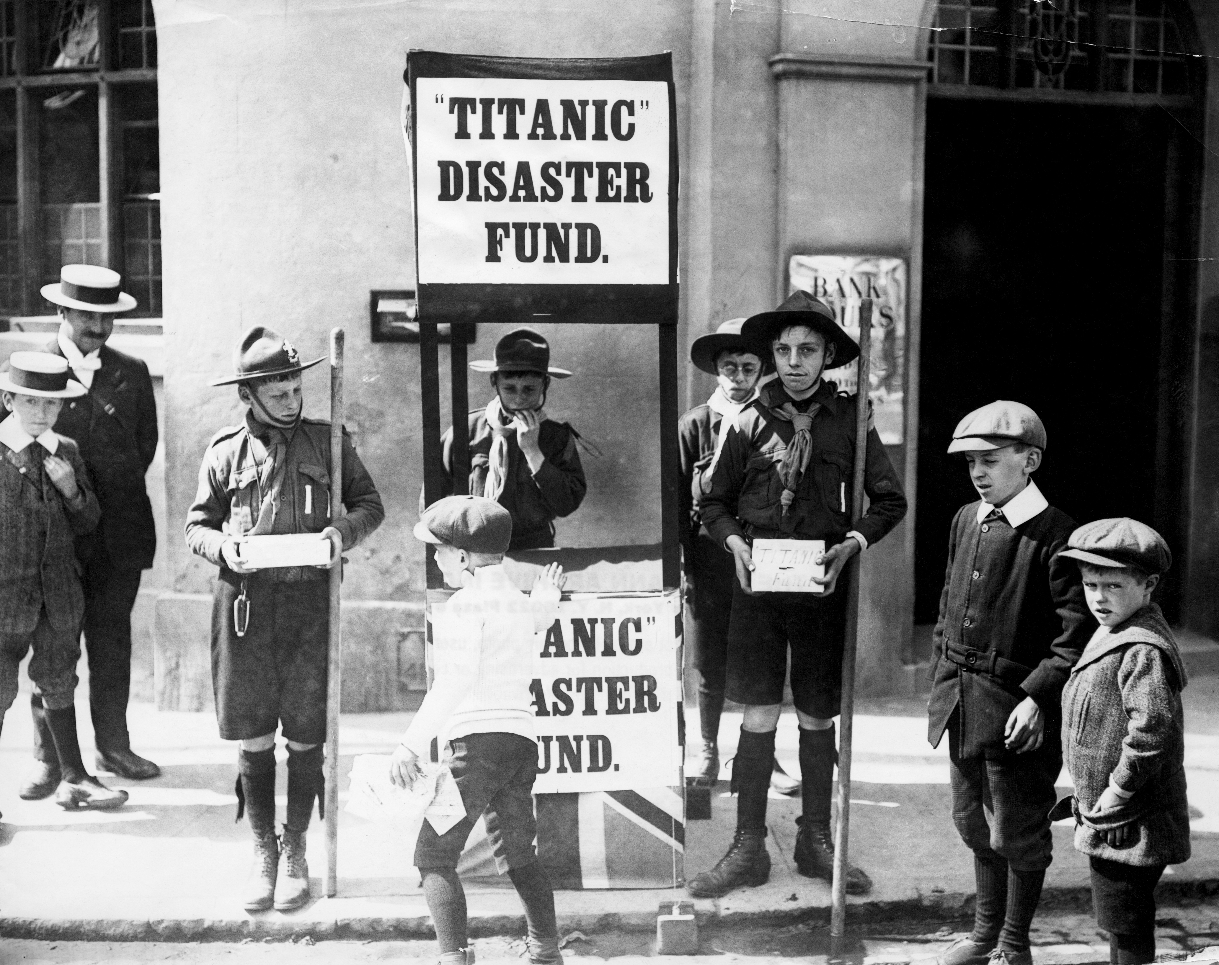 Children collecting donations for the Titanic disaster fund, standing with donation boxes and a sign outside a building