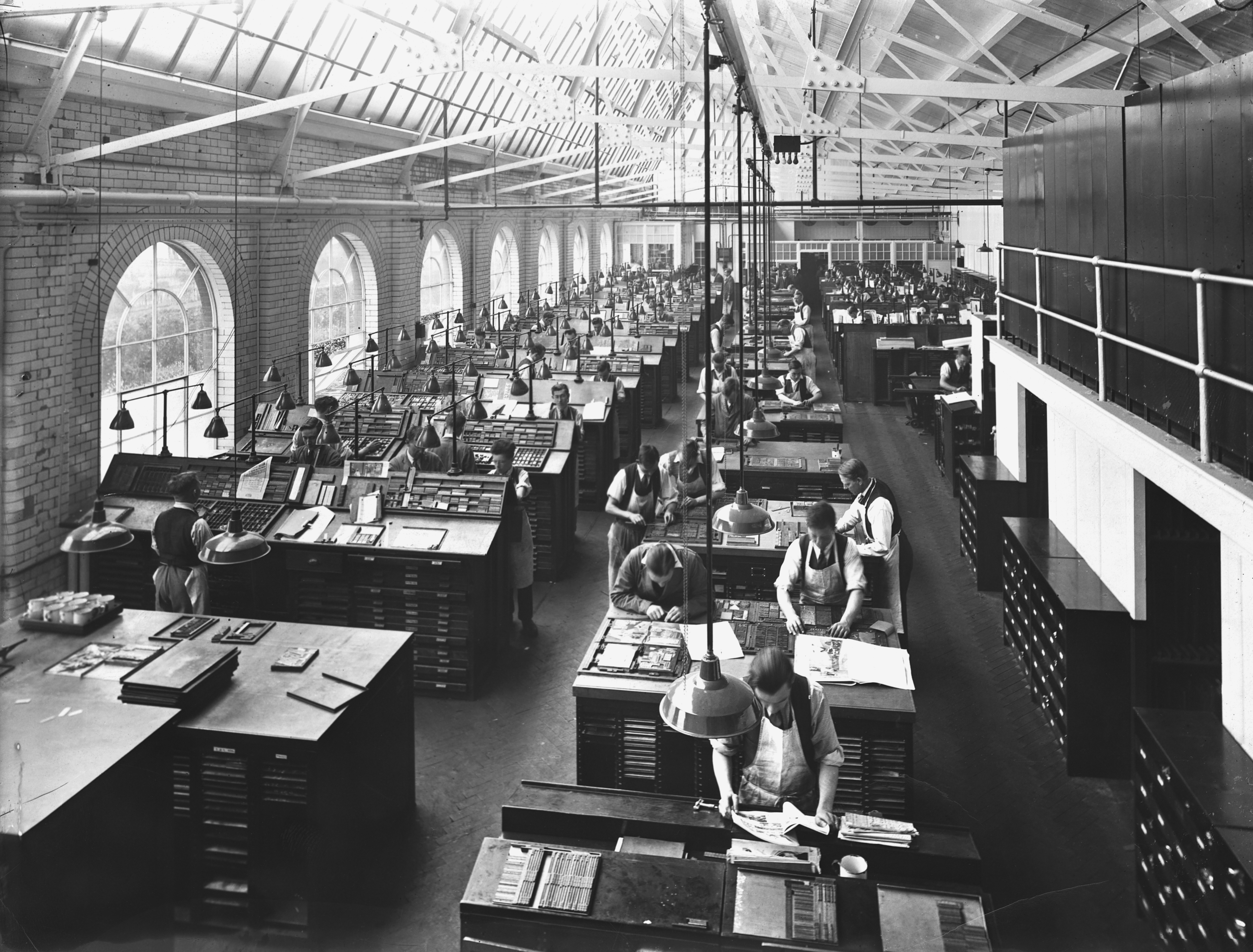 Historic printing press room with workers operating machines and arranging type, beneath large arched windows and high ceiling
