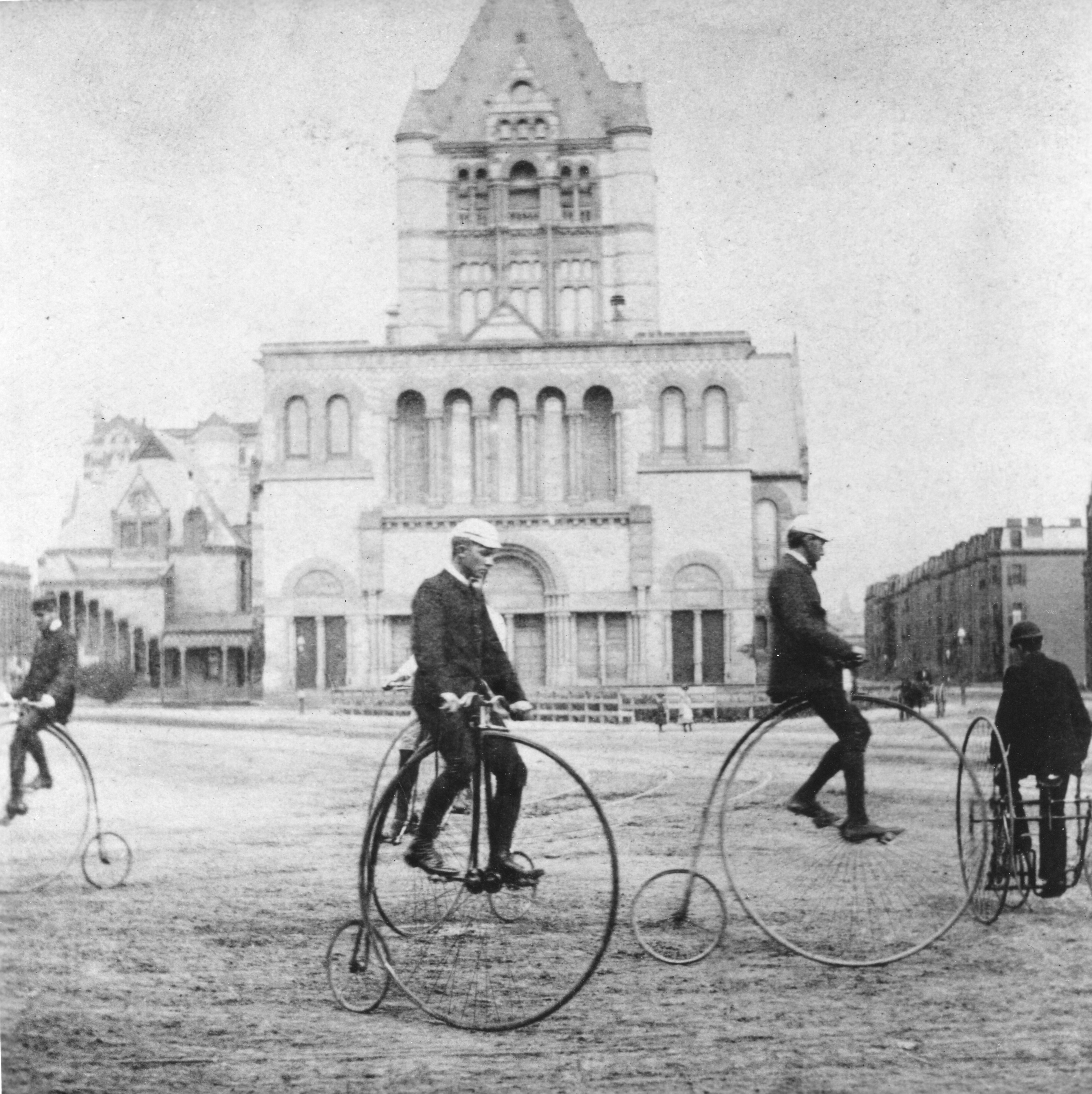 People riding penny-farthing bicycles in a historical setting, with an ornate building in the background