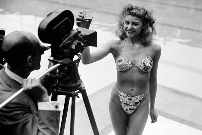 A woman in a patterned bikini poses confidently for a photographer with a large camera near a swimming pool