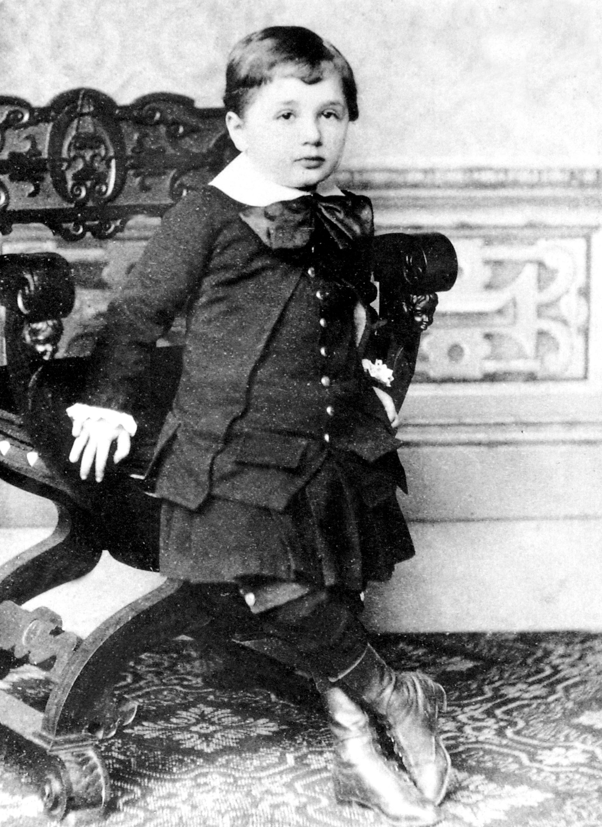 Young child in formal early 20th-century attire, sitting on ornately carved chair, looking at camera