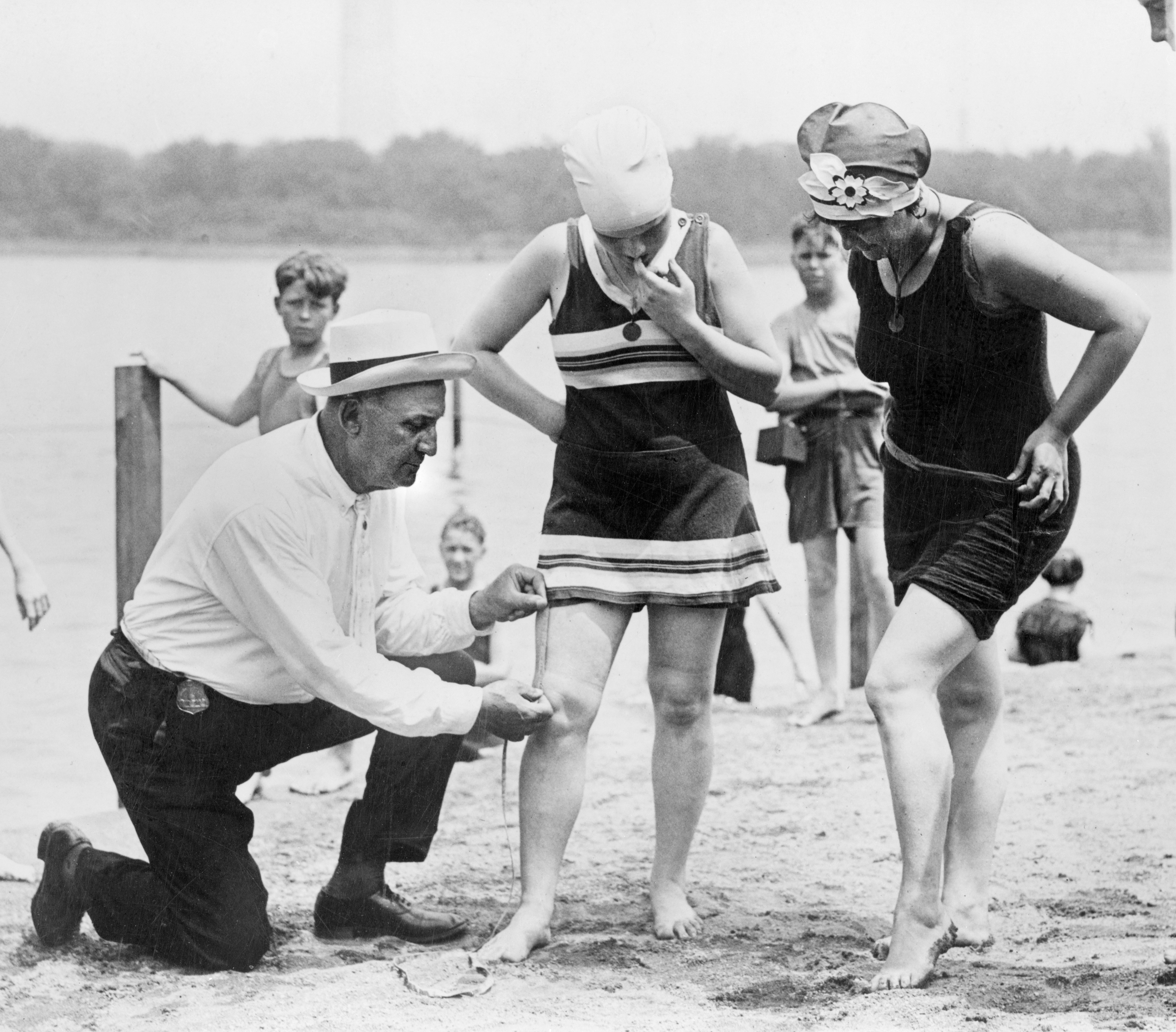 A man measures a woman's swimsuit length with a tape measure on a beach as other beachgoers look on