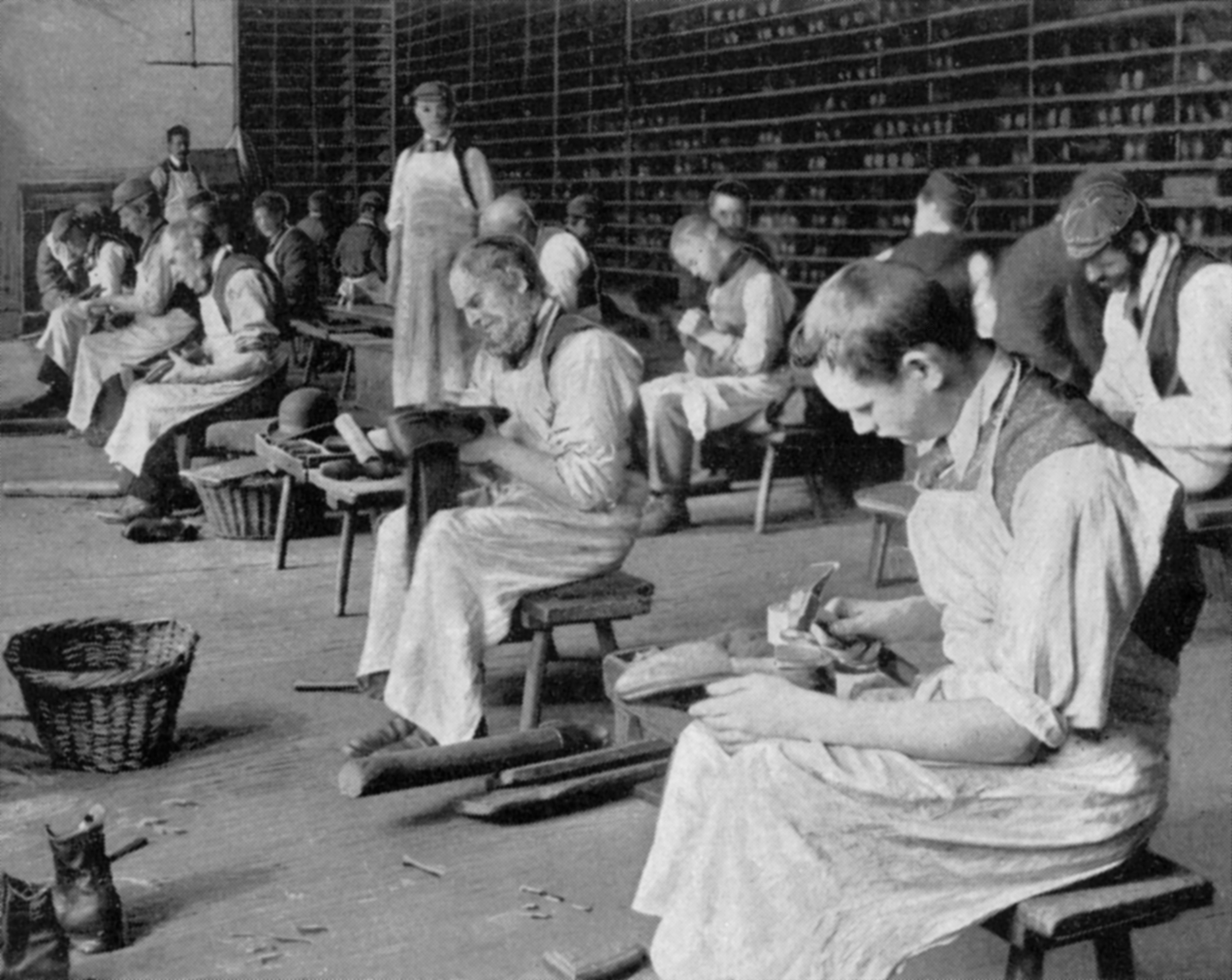 Workers in aprons craft shoes in a vintage cobbler workshop. Shelves filled with materials line the walls