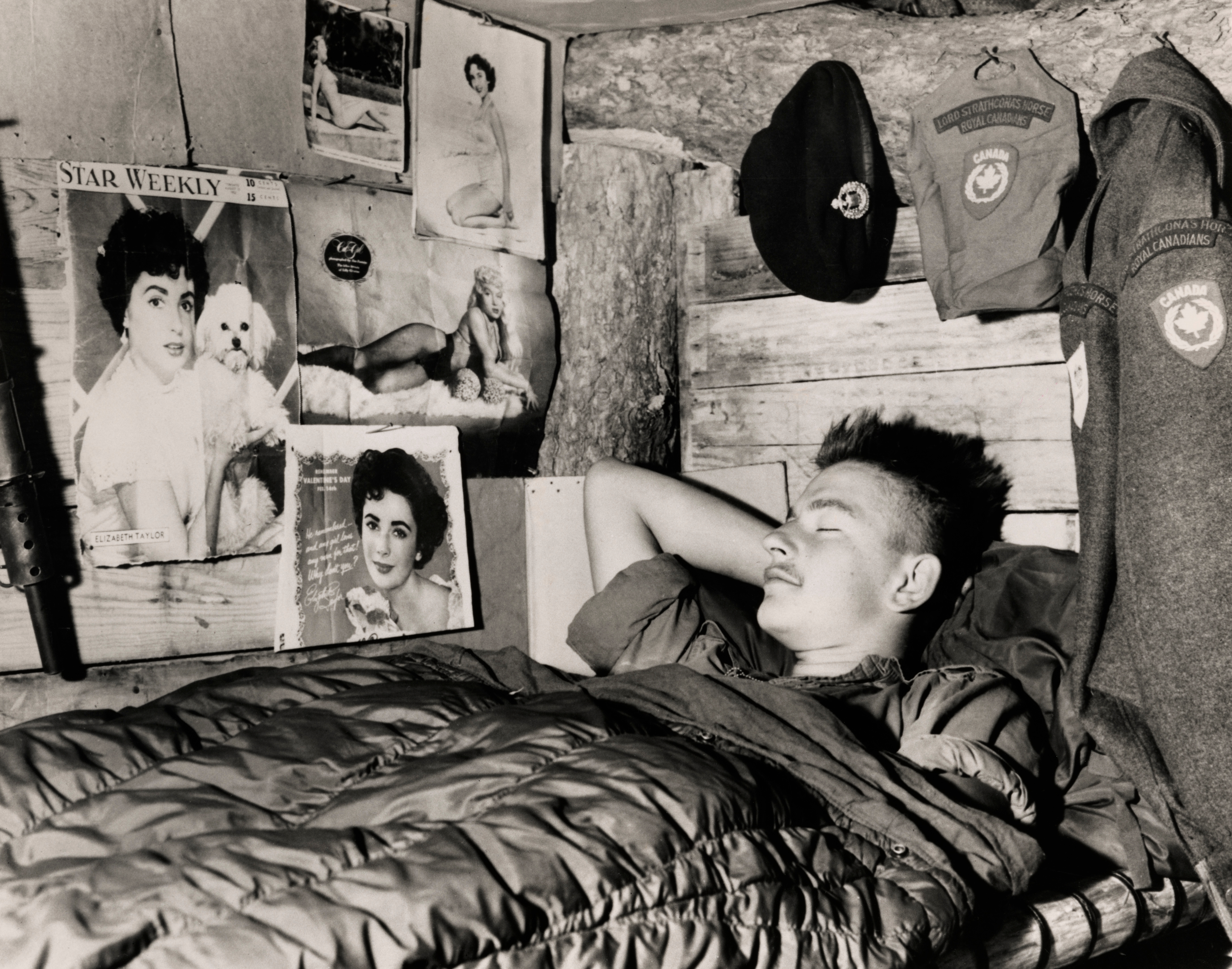 A soldier sleeps in a cabin decorated with celebrity posters and a uniform jacket hangs nearby