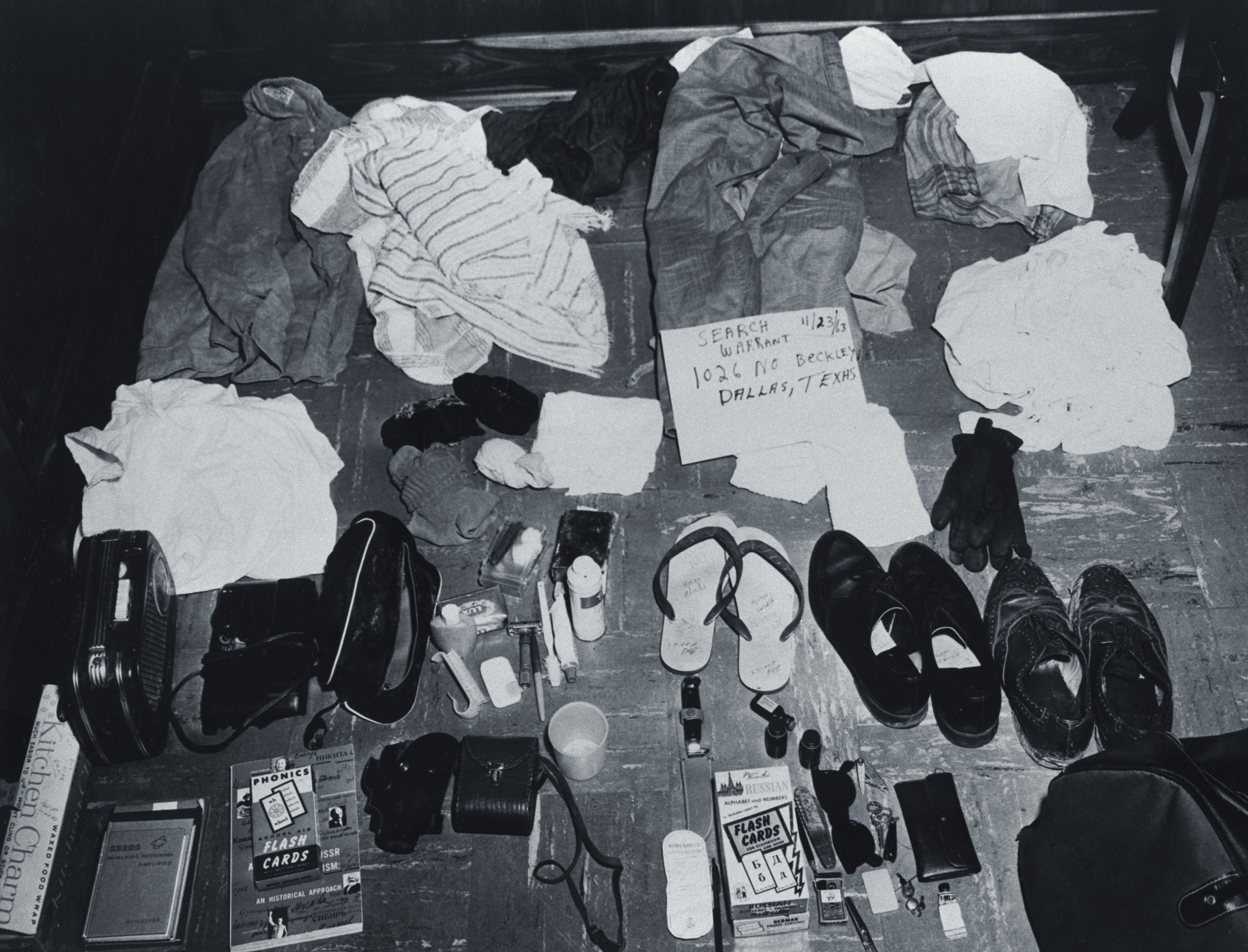 Clothing, shoes, toiletries, and personal items are laid out neatly on the floor, along with a note reading "Search 1/64, 1026 North Beckley, Dallas, Texas."