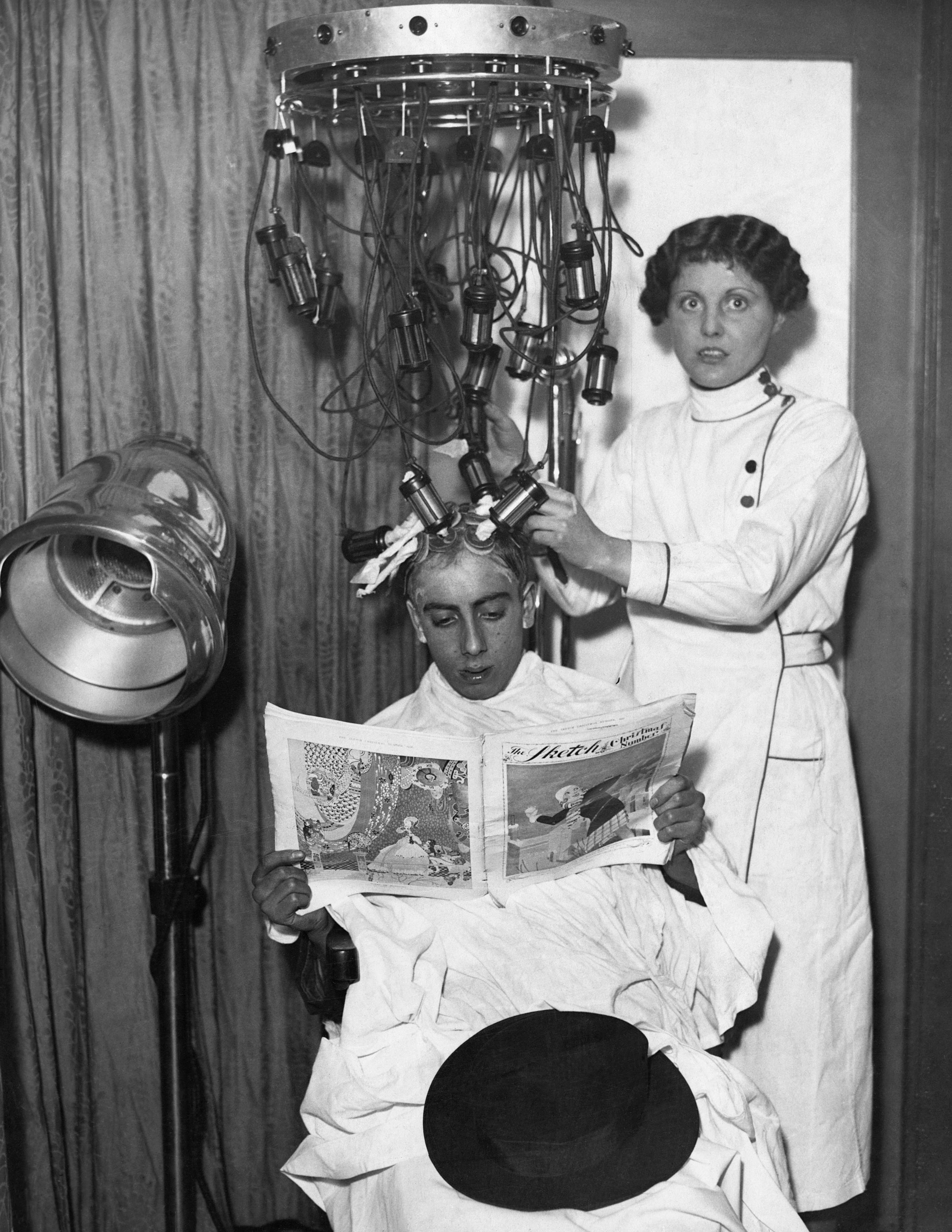 Person seated, reading a newspaper under a vintage hair perm machine, attended by a stylist in a white uniform
