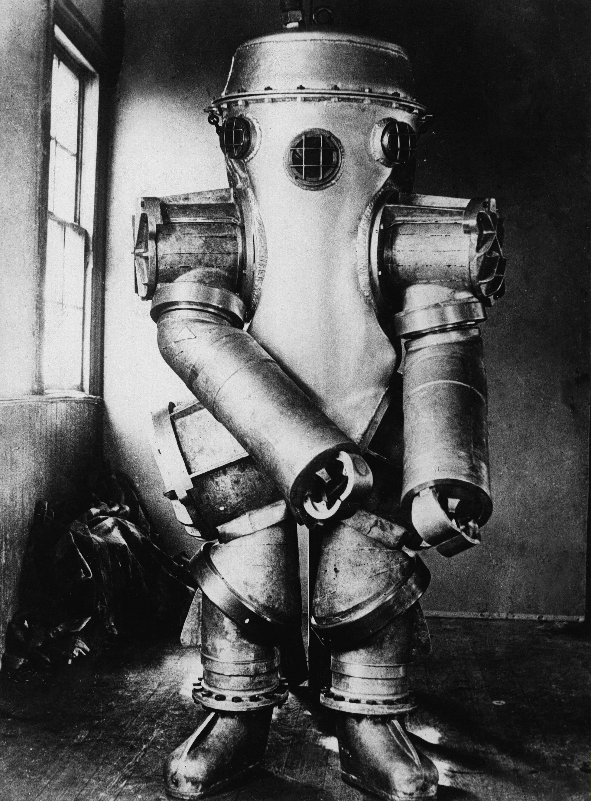 Vintage deep-sea diving suit with metal exterior and round helmet, resembling a robot. Standing indoors near a window