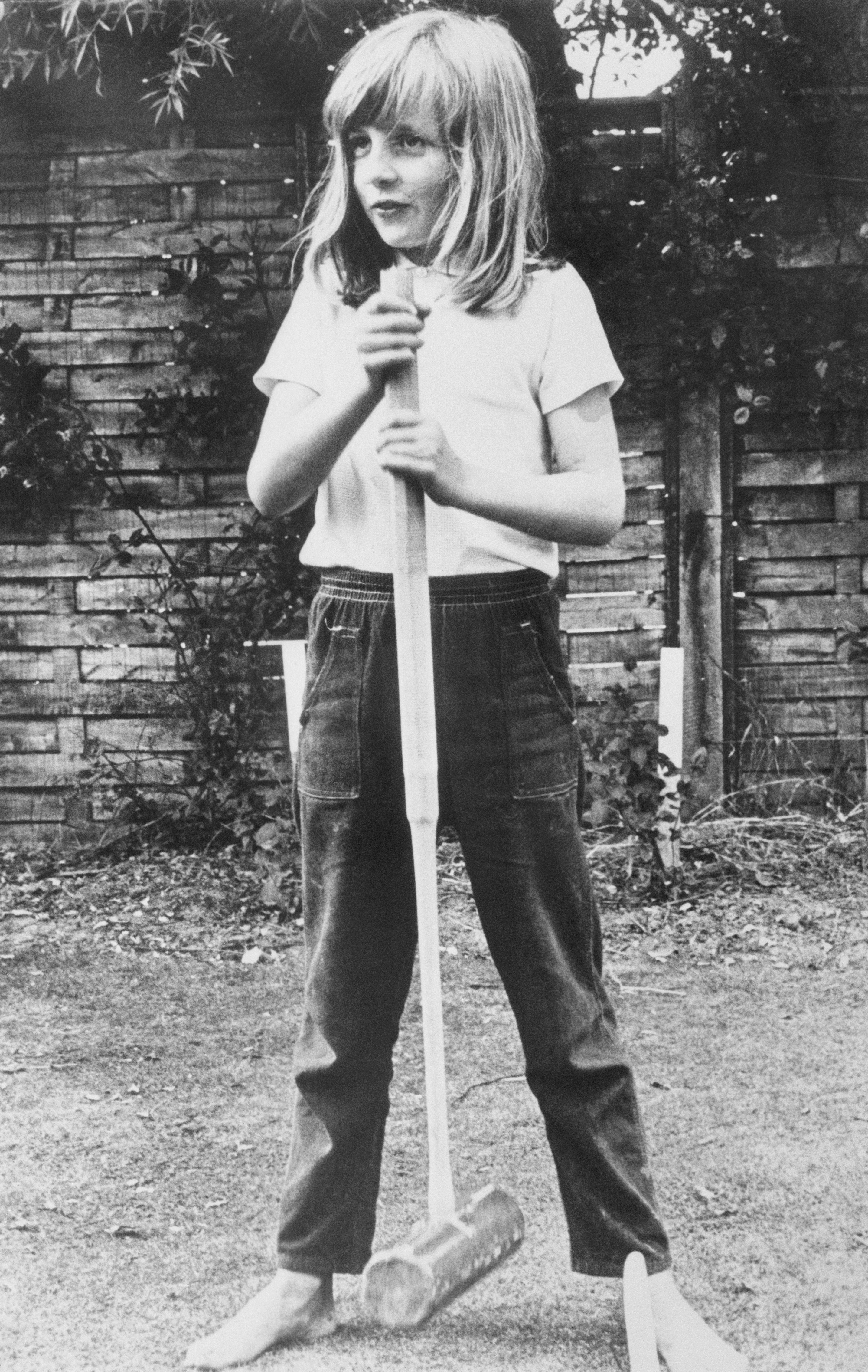 Child playing croquet barefoot on grass, holding a mallet. They have long hair and are wearing a t-shirt and jeans. Background shows a garden fence
