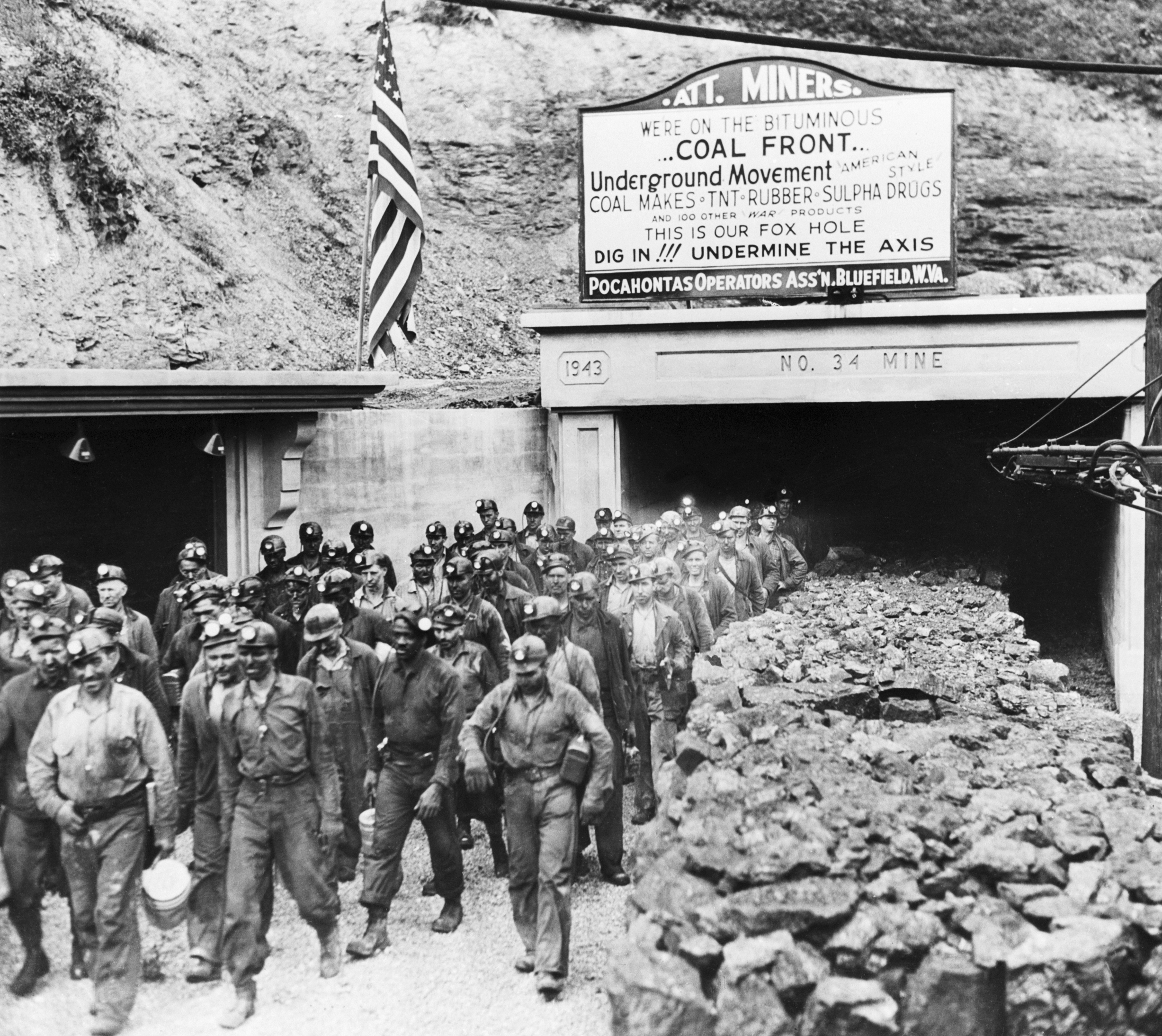 Miners exit a coal mine in 1943, wearing helmets and work clothes. A sign above the entrance promotes coal for the war effort against the Axis