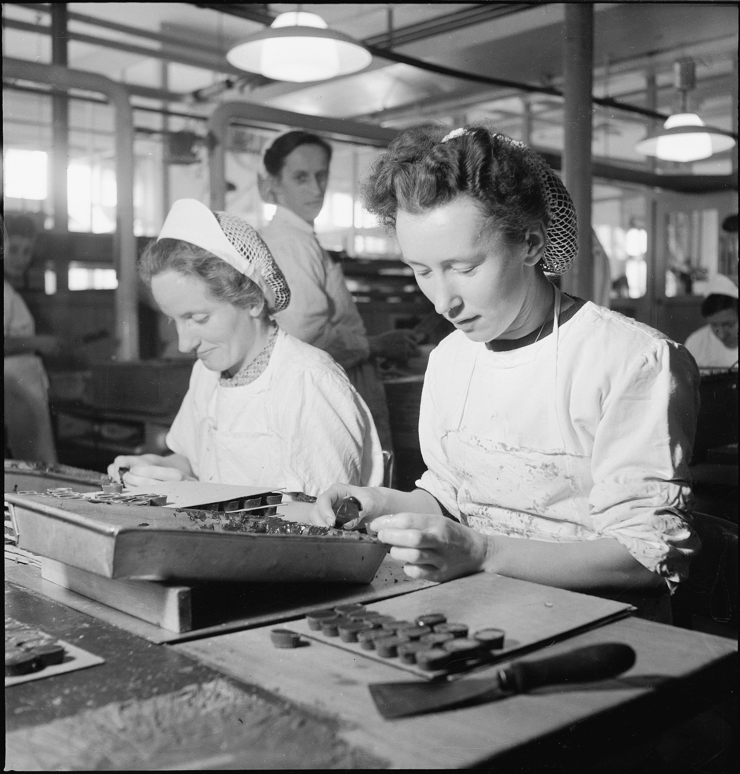 Two women are working at a chocolate factory, wearing aprons and hairnets, focusing on arranging chocolates on trays