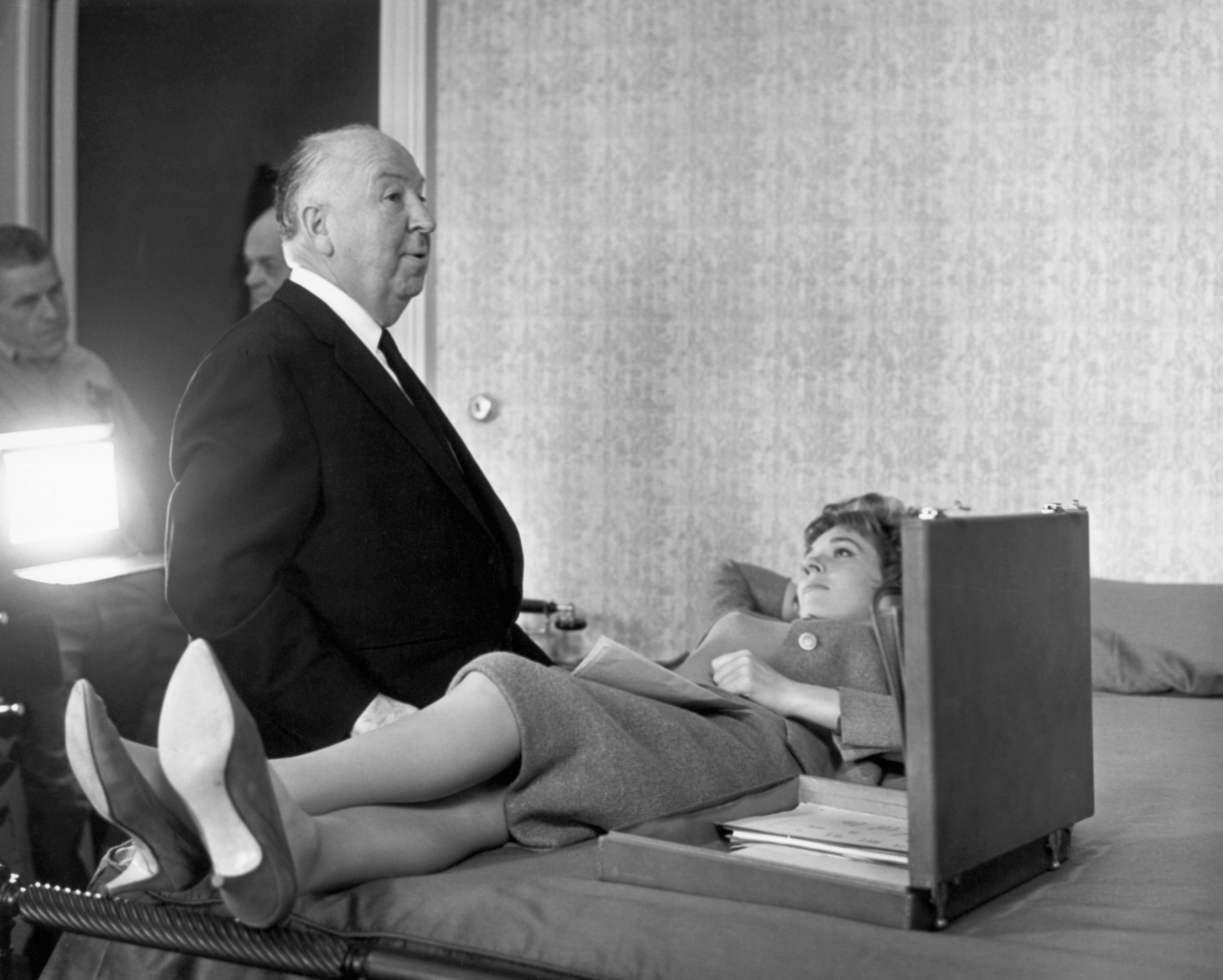 A director and an actress on a set; the actress appears to be lying on a bed, suggesting a film scene. Nearby crew members observe