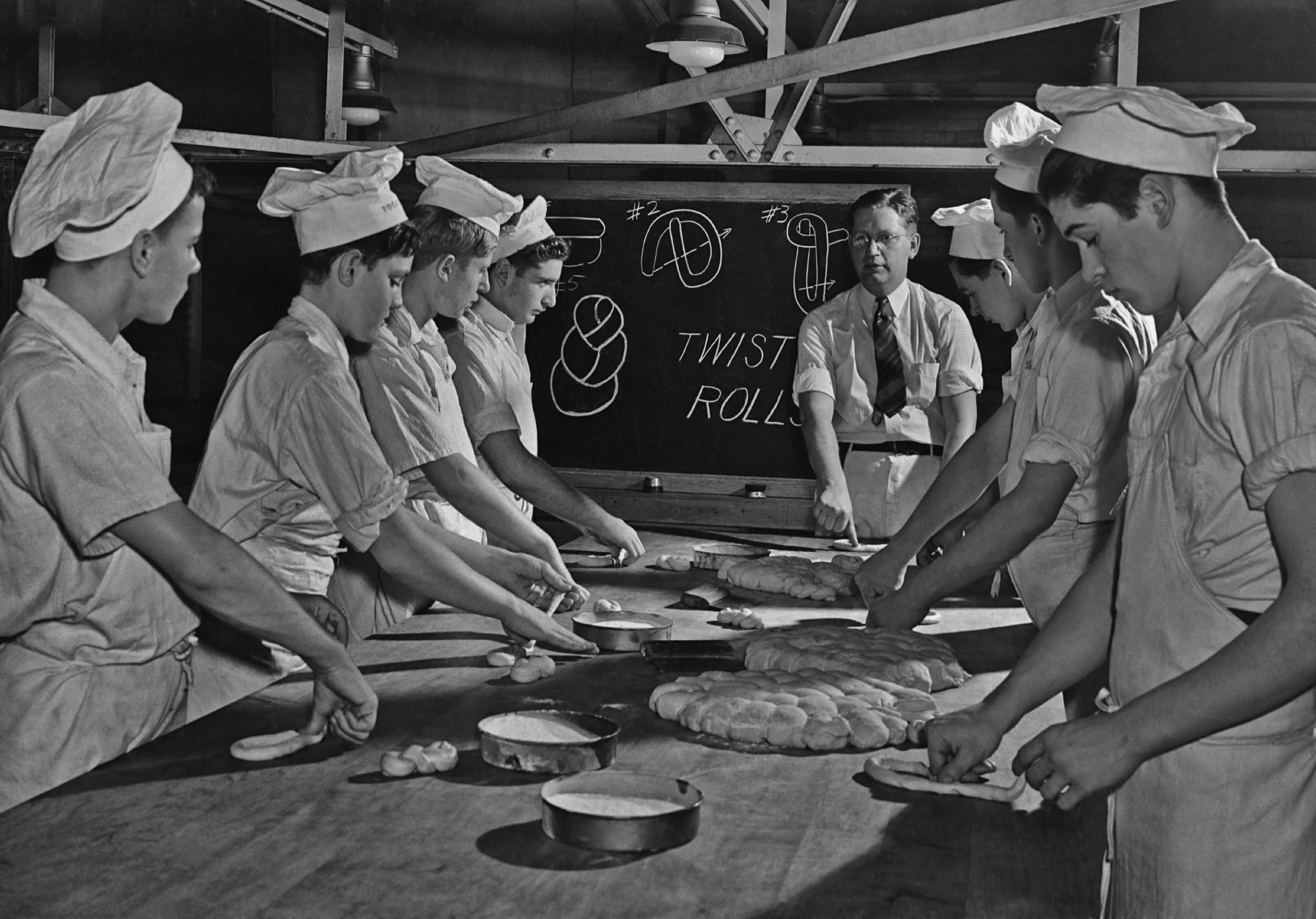 Bakers in uniform learn to shape dough into twisted rolls in a bakery class, guided by an instructor