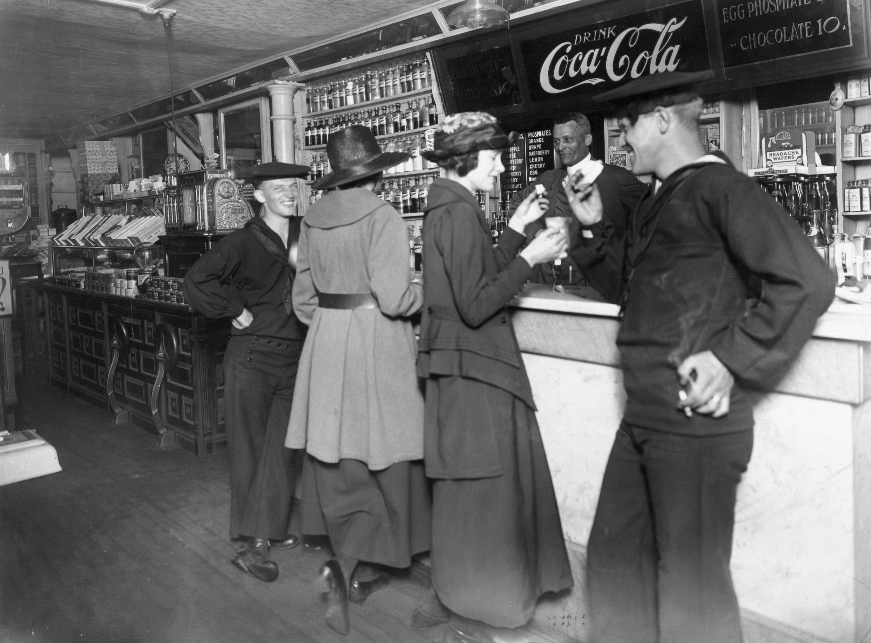 People in early 20th-century attire enjoy drinks at a soda fountain counter, with a Coca-Cola sign in the background
