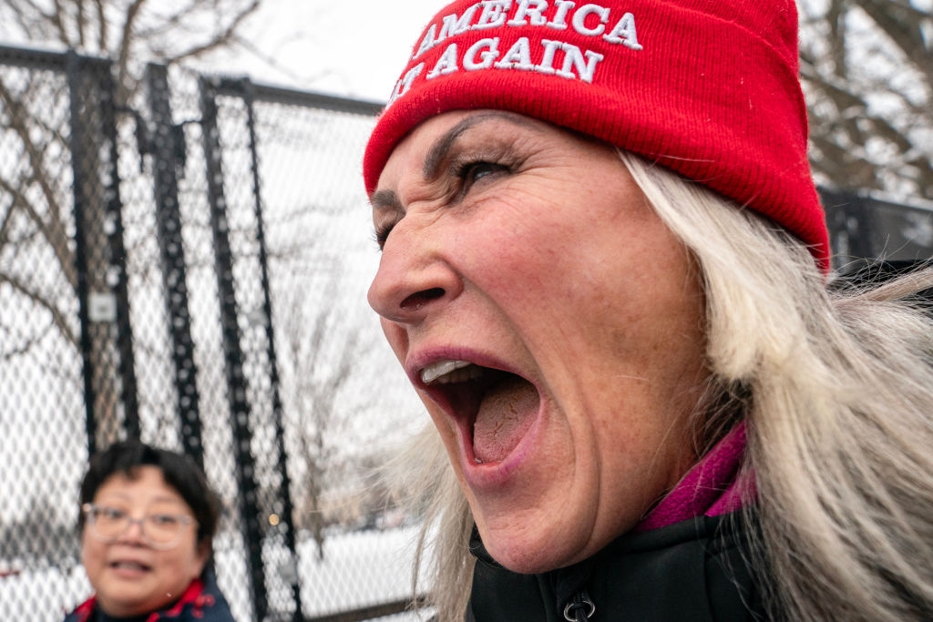 A woman wearing a "Make America Great Again" hat shouts passionately, while another person stands behind her near a fence