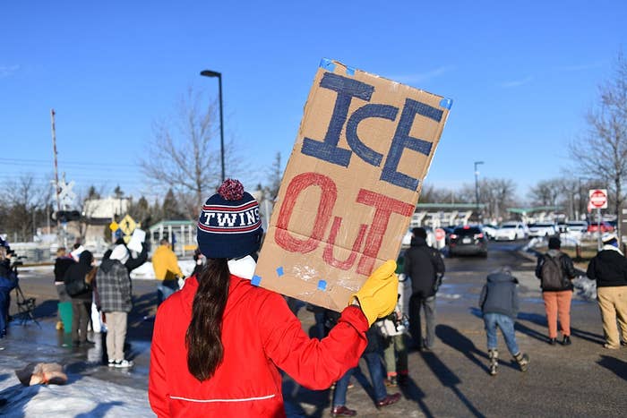 Person holding "Ice Out" sign at rally, with others gathered in a snowy area, possibly protesting immigration policies