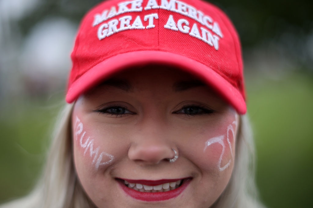 Person wearing a "Make America Great Again" cap with "Trump 2020" written on their cheeks in a park setting