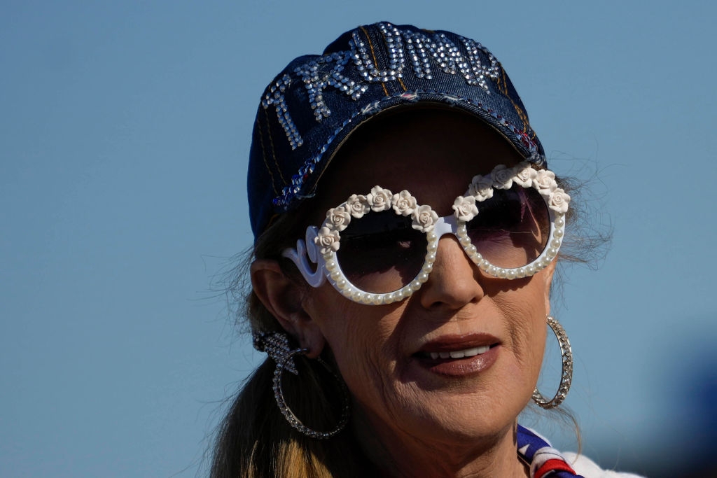 Person wearing a cap with rhinestone lettering and floral sunglasses, large hoop earrings, speaking outside